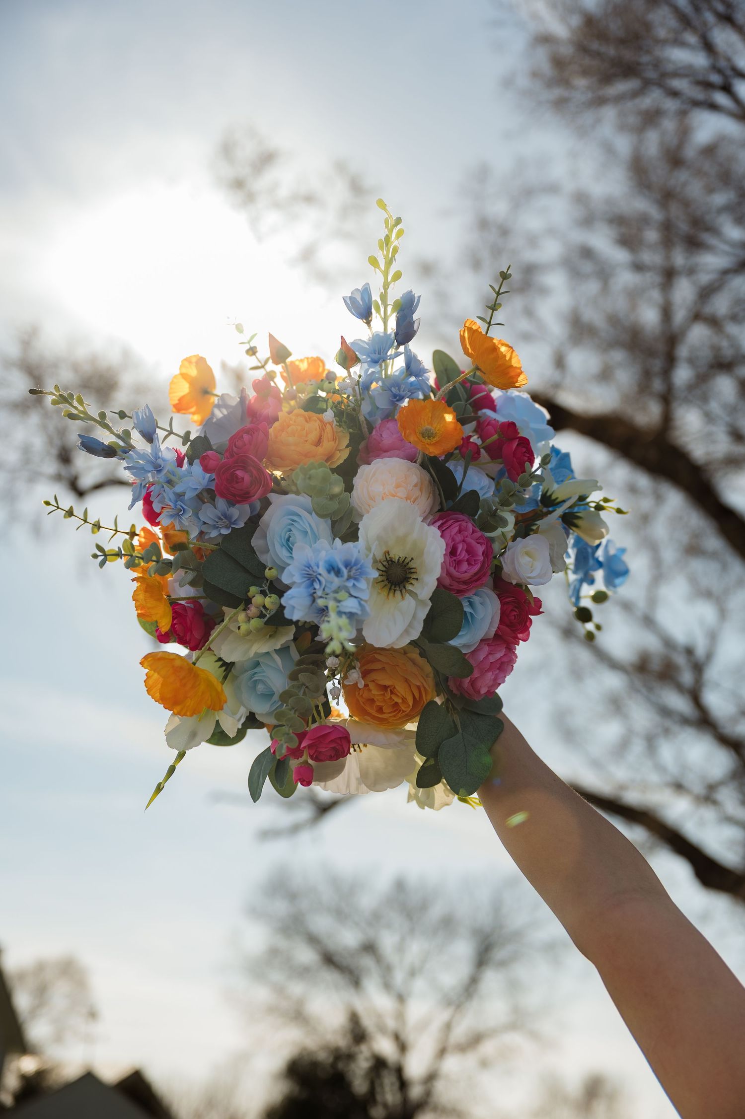 A vibrant bouquet of mixed flowers held up against a bright sunlit sky, featuring orange, blue, and pink blooms.