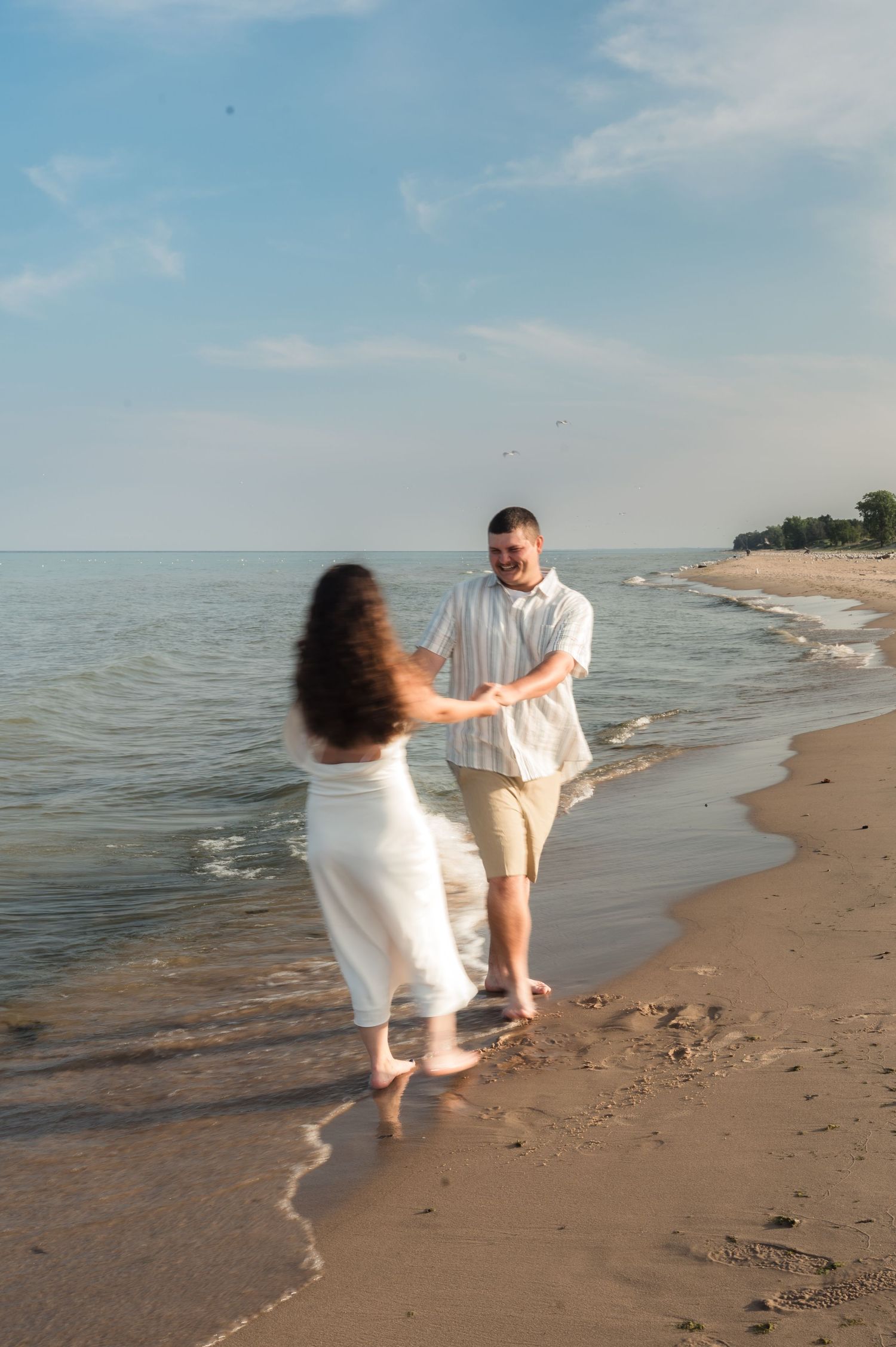 A playful couple in white attire runs and dances along a serene beach shoreline on a sunny day.