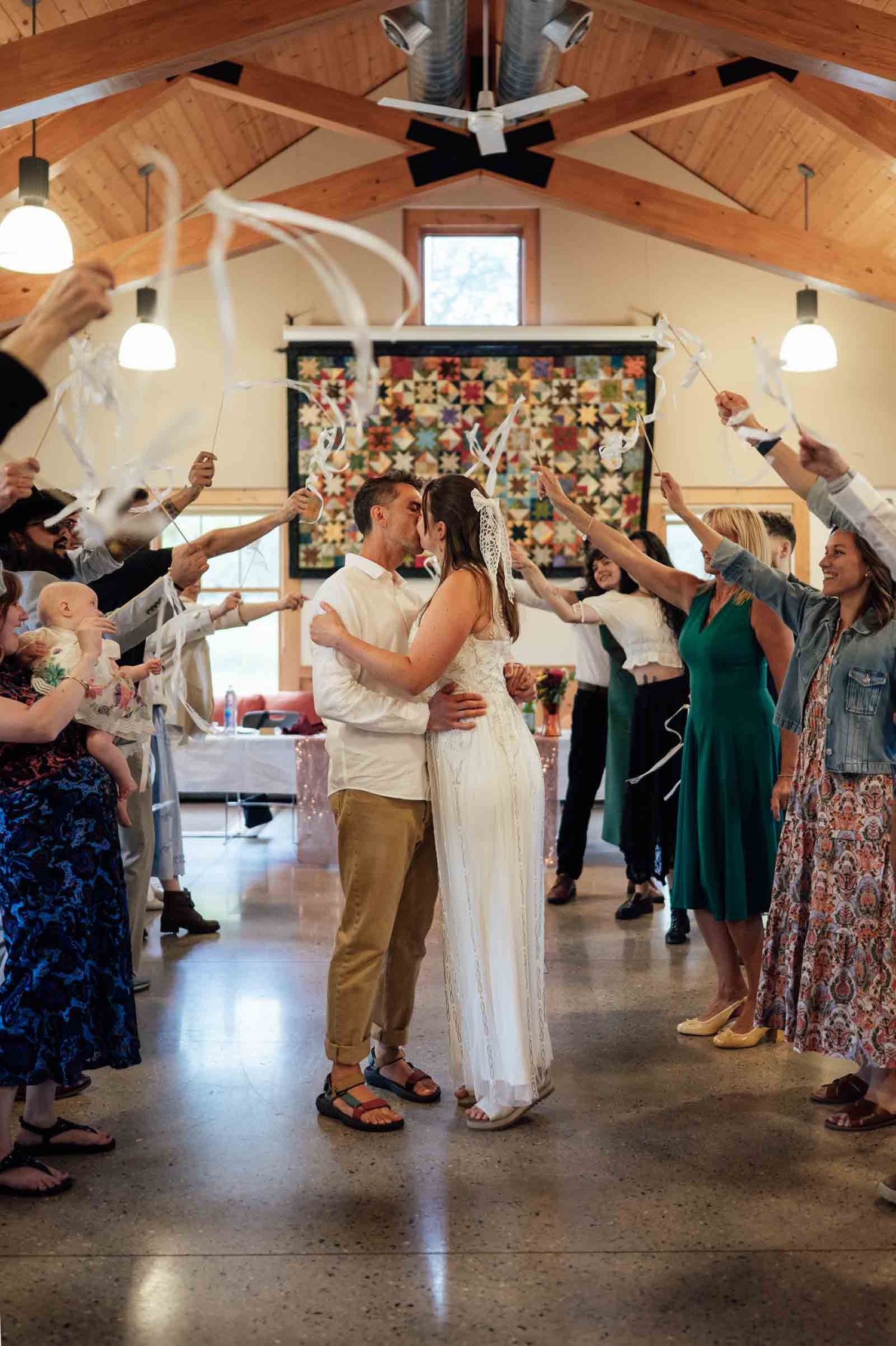 Newlyweds share a kiss under a ribbon arch surrounded by cheering wedding guests in a rustic venue.
