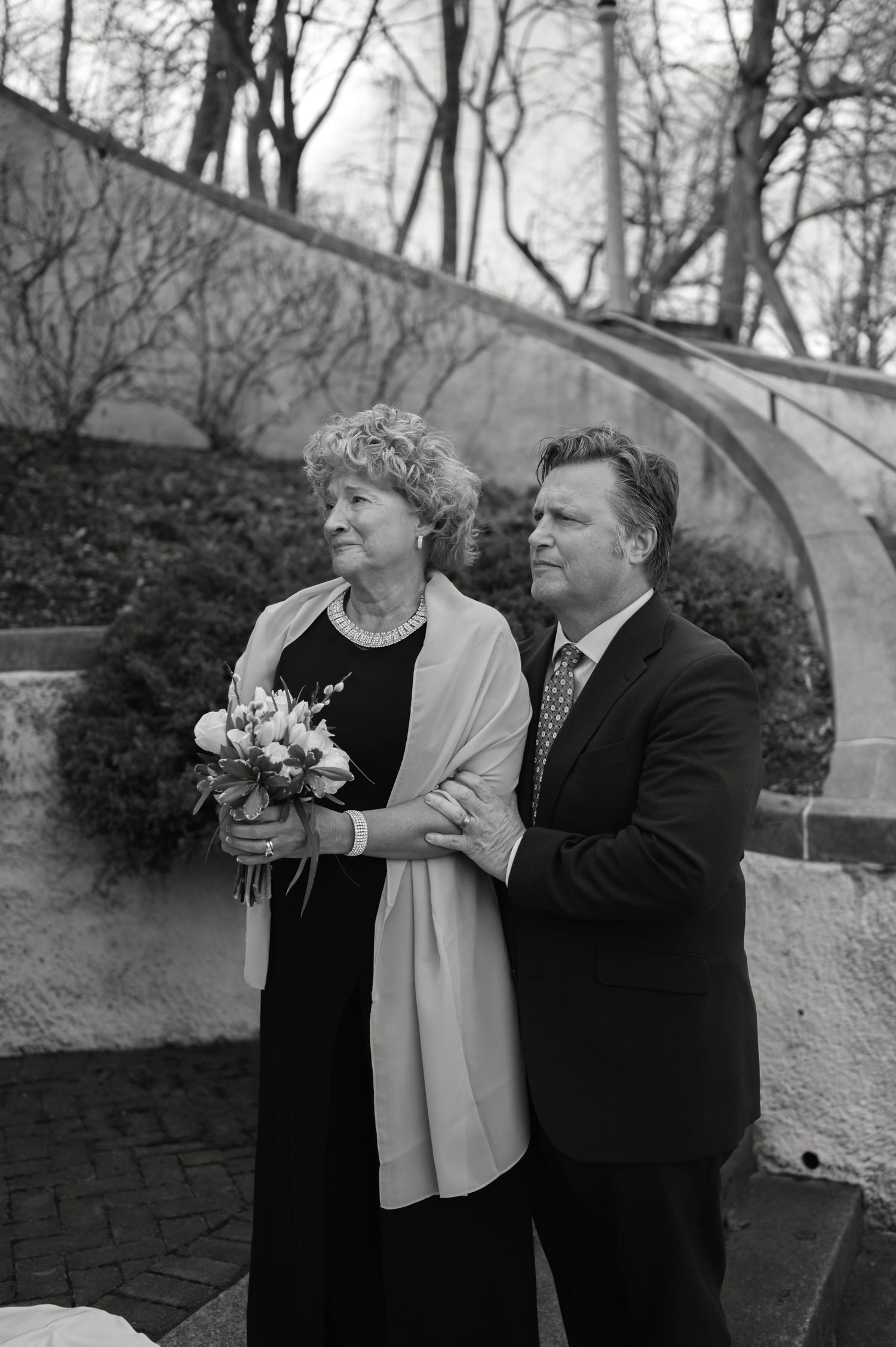Elegant couple in formal attire holds bouquet of flowers outdoors in black and white photo.