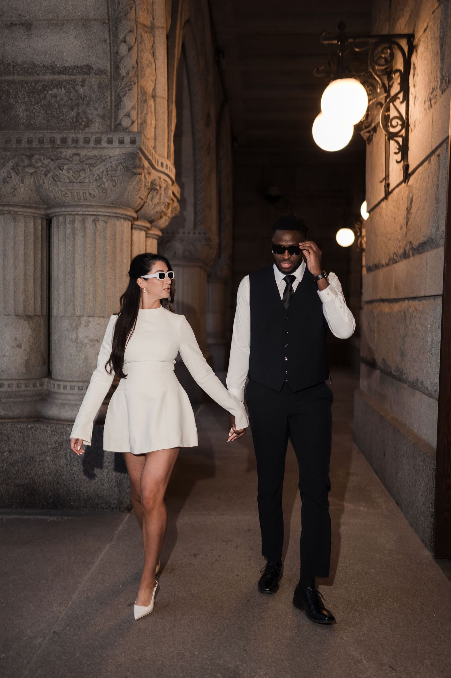 Elegant couple in white dress and black suit walking hand-in-hand through a grand lit colonnade.