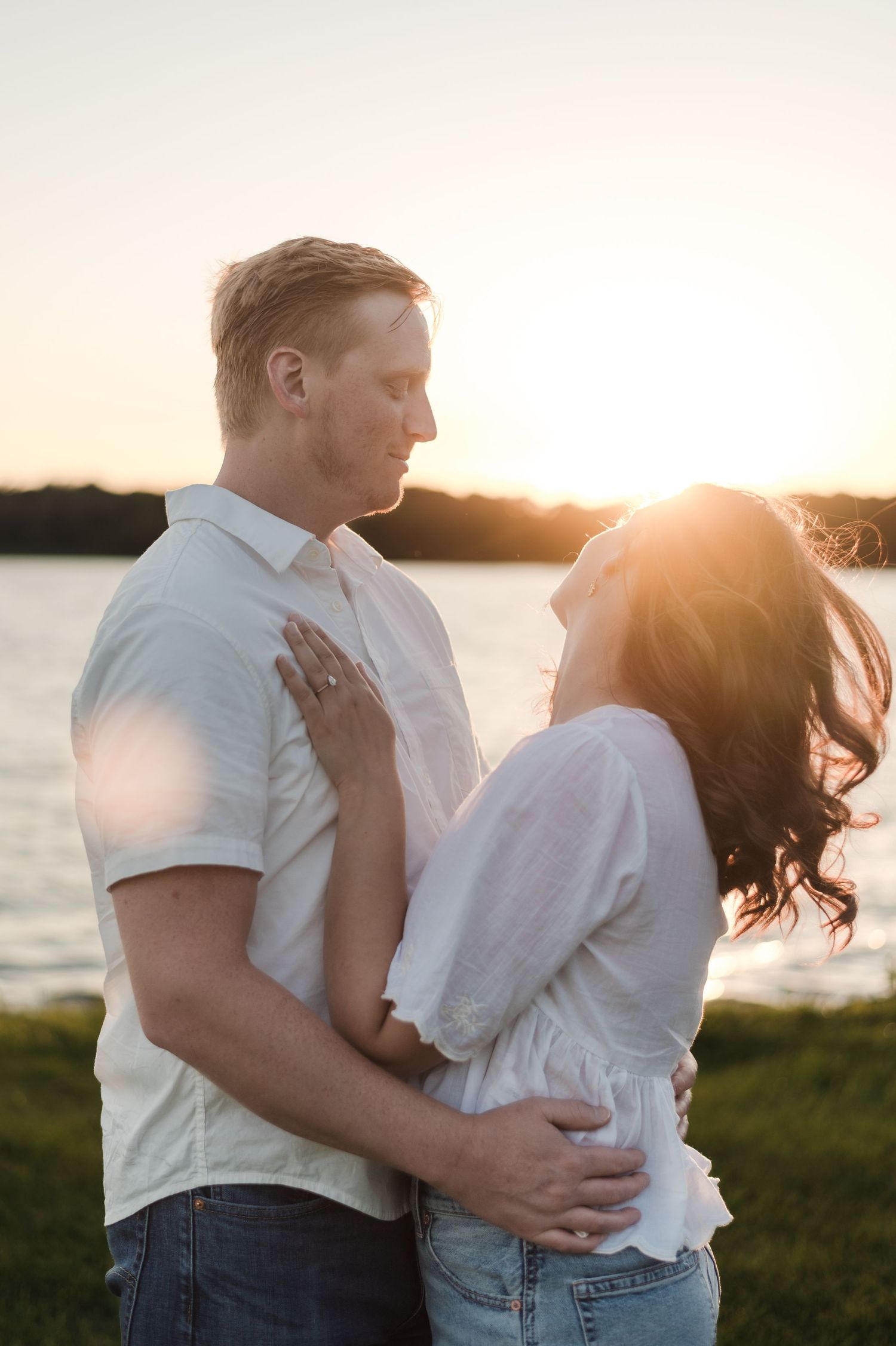Romantic couple embraces near a lake at sunset, woman showing engagement ring on mans chest.
