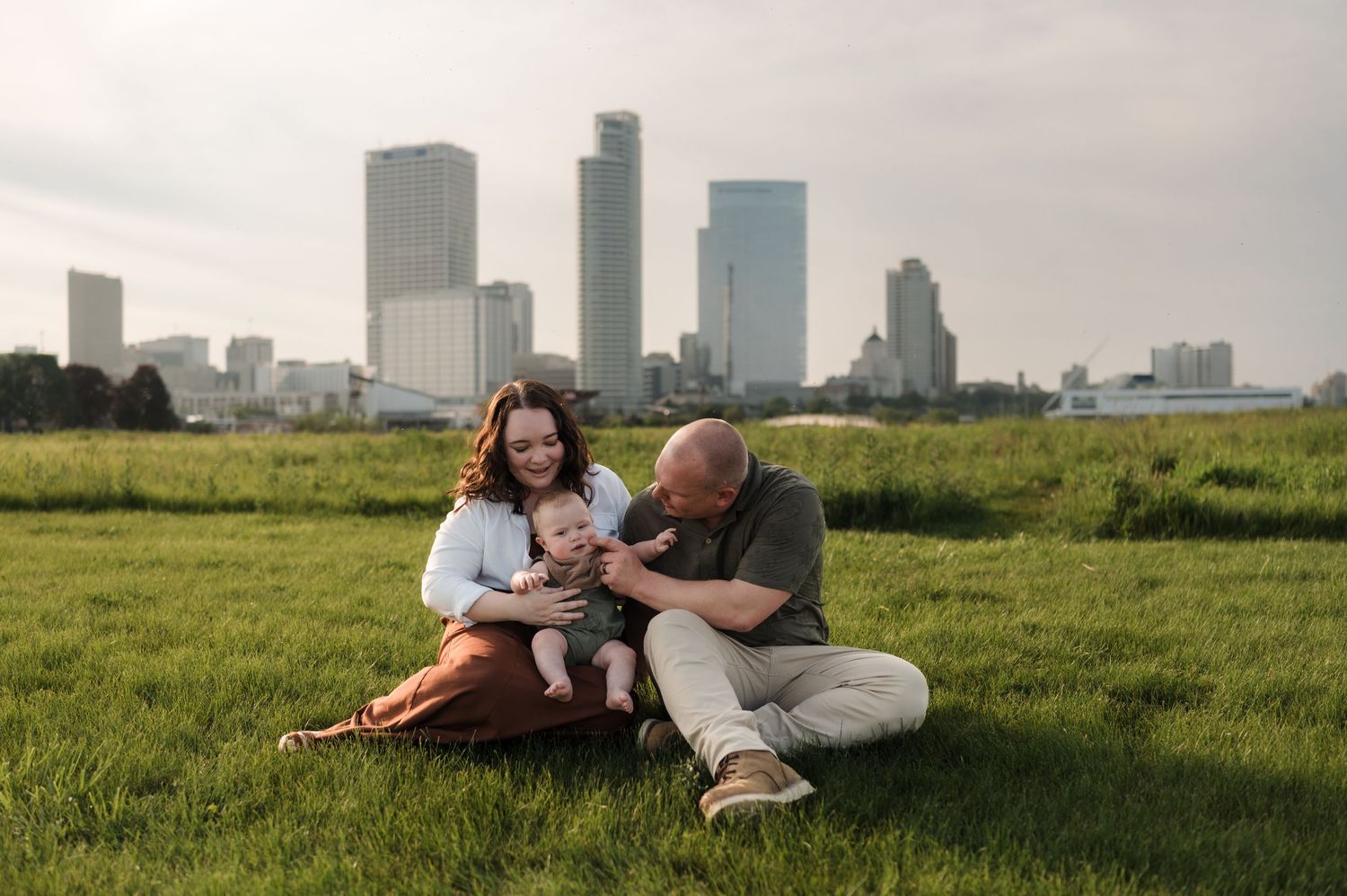 Happy family of three sitting together on green grass with a city skyline at golden hour.