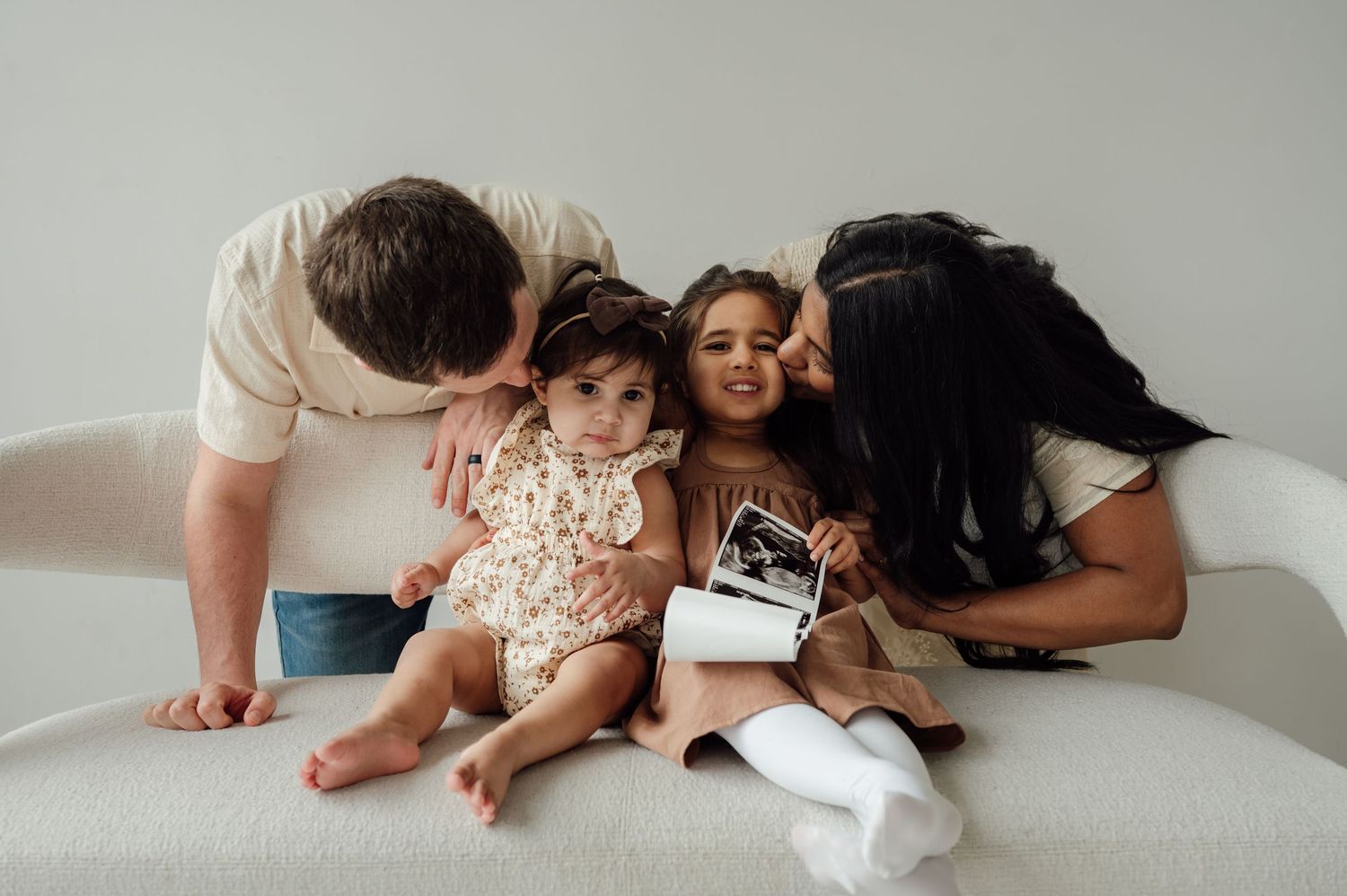 Parents kissing two young daughters holding ultrasound photos on white sofa during pregnancy reveal.
