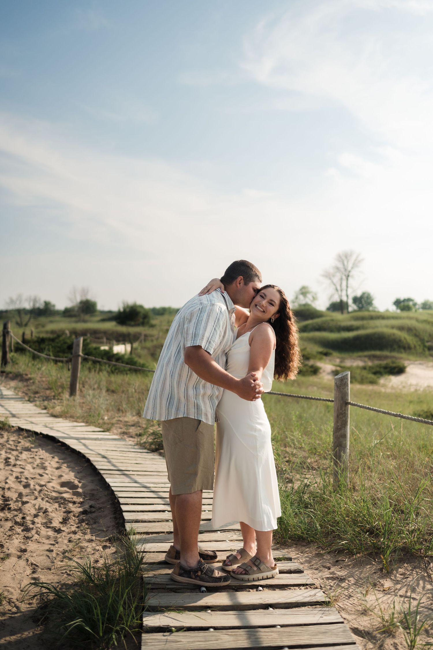 A couple share an intimate moment on a wooden boardwalk path along sandy beach dunes during golden hour.