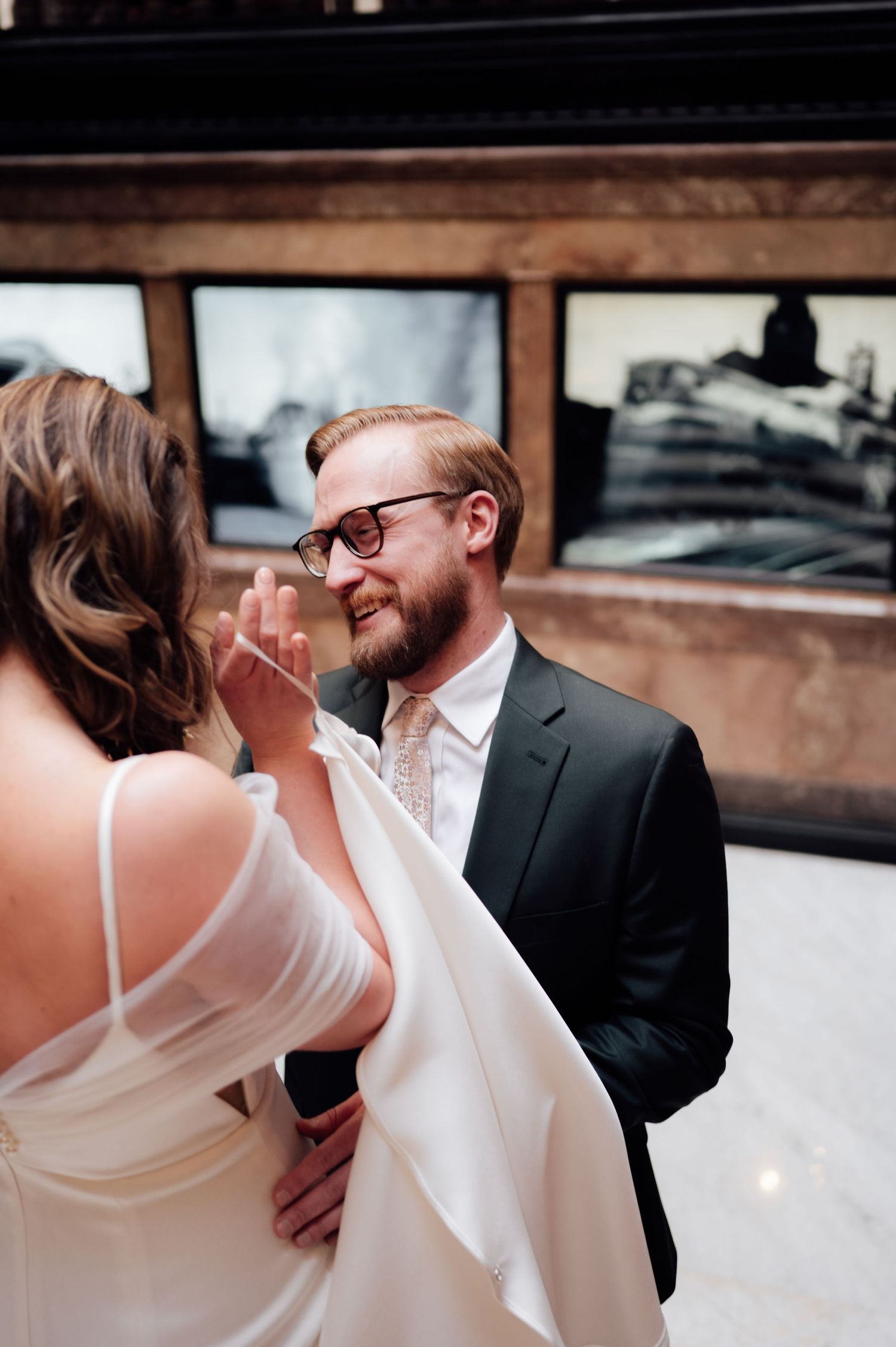 Smiling groom in black suit and glasses holds brides hand during elegant indoor wedding ceremony.