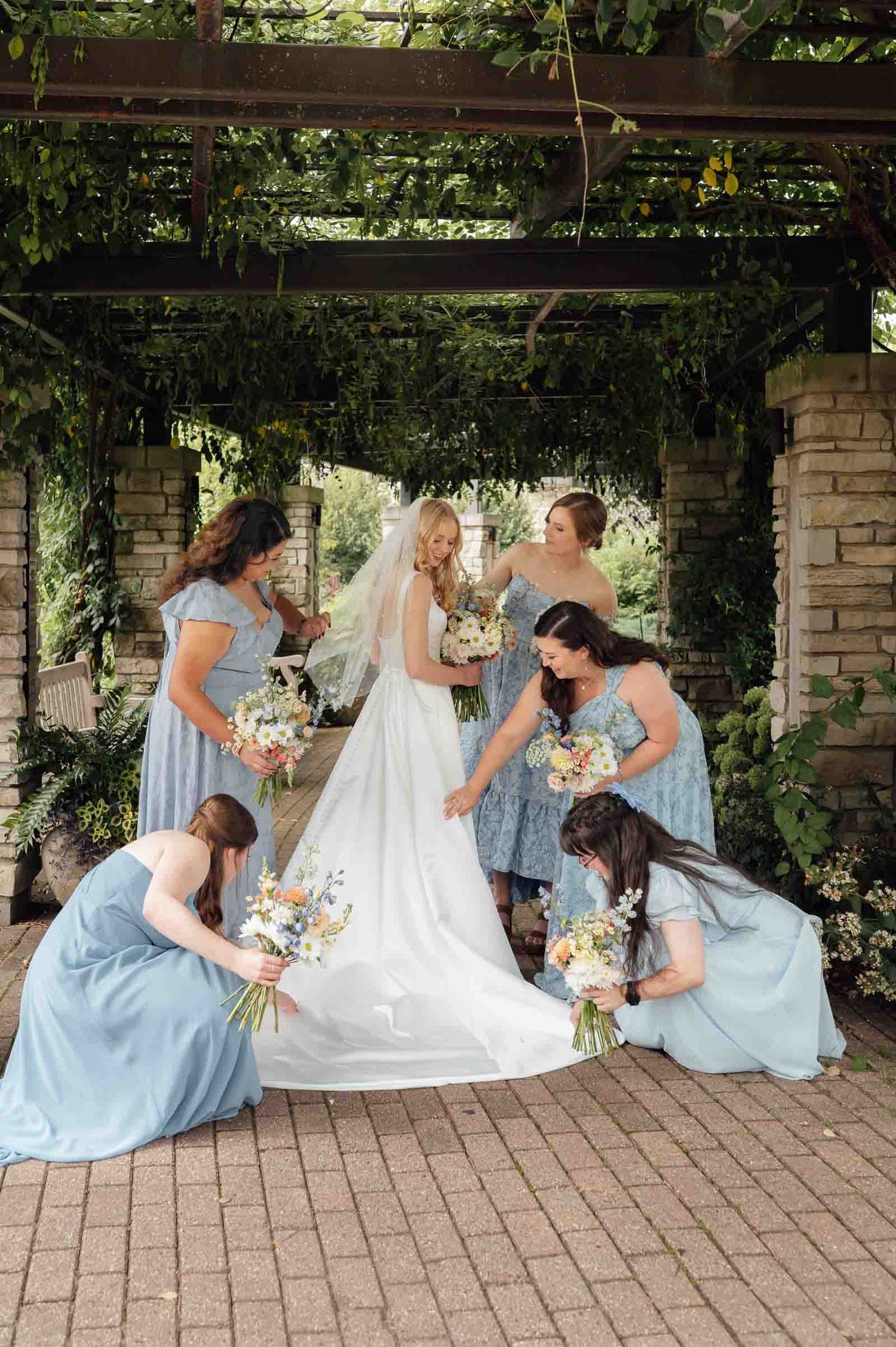 Bridesmaids in blue dresses arrange bride's veil and train under vine-covered stone pergola.