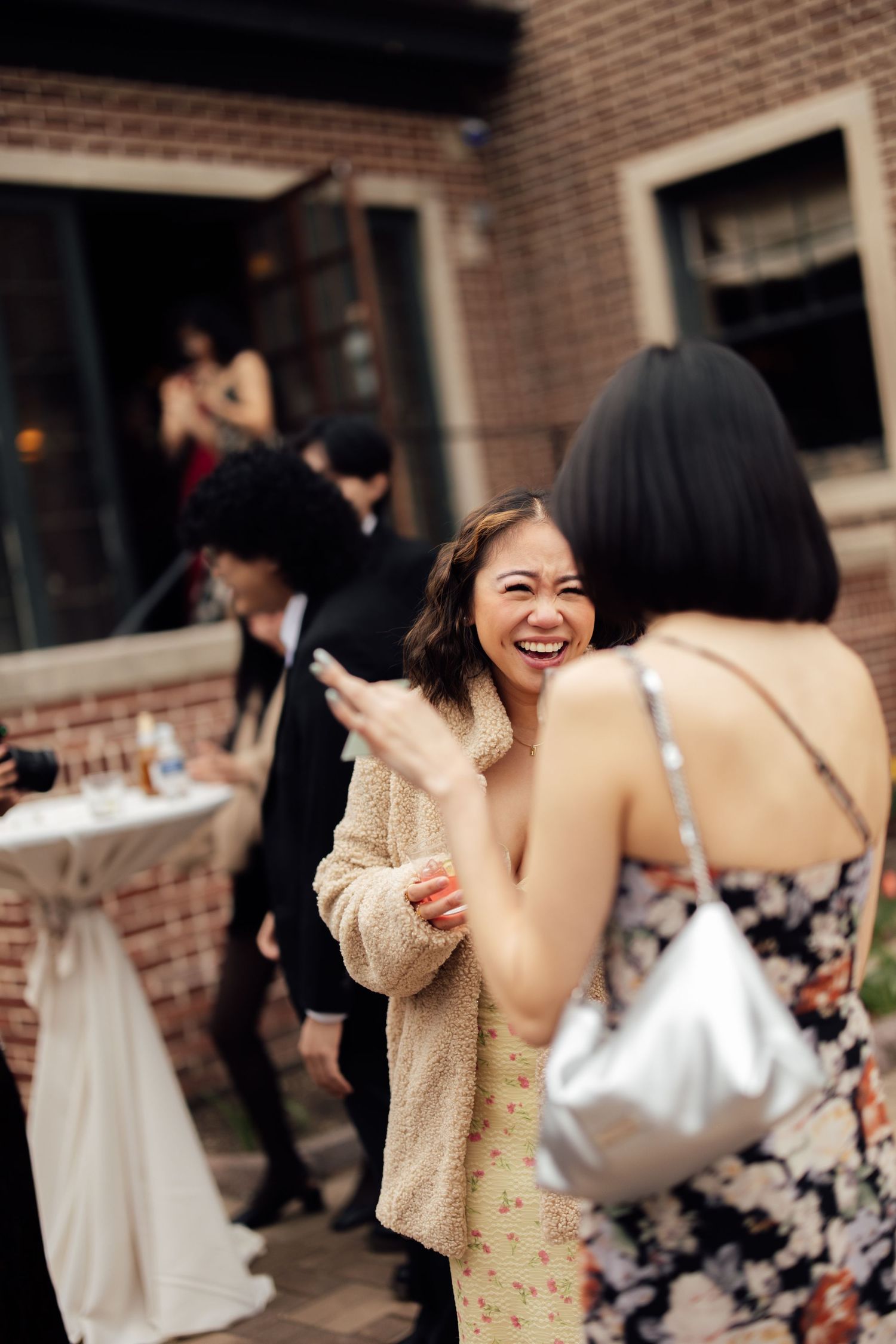 Wedding guest in floral dress and teddy coat laughs joyfully at outdoor reception gathering.