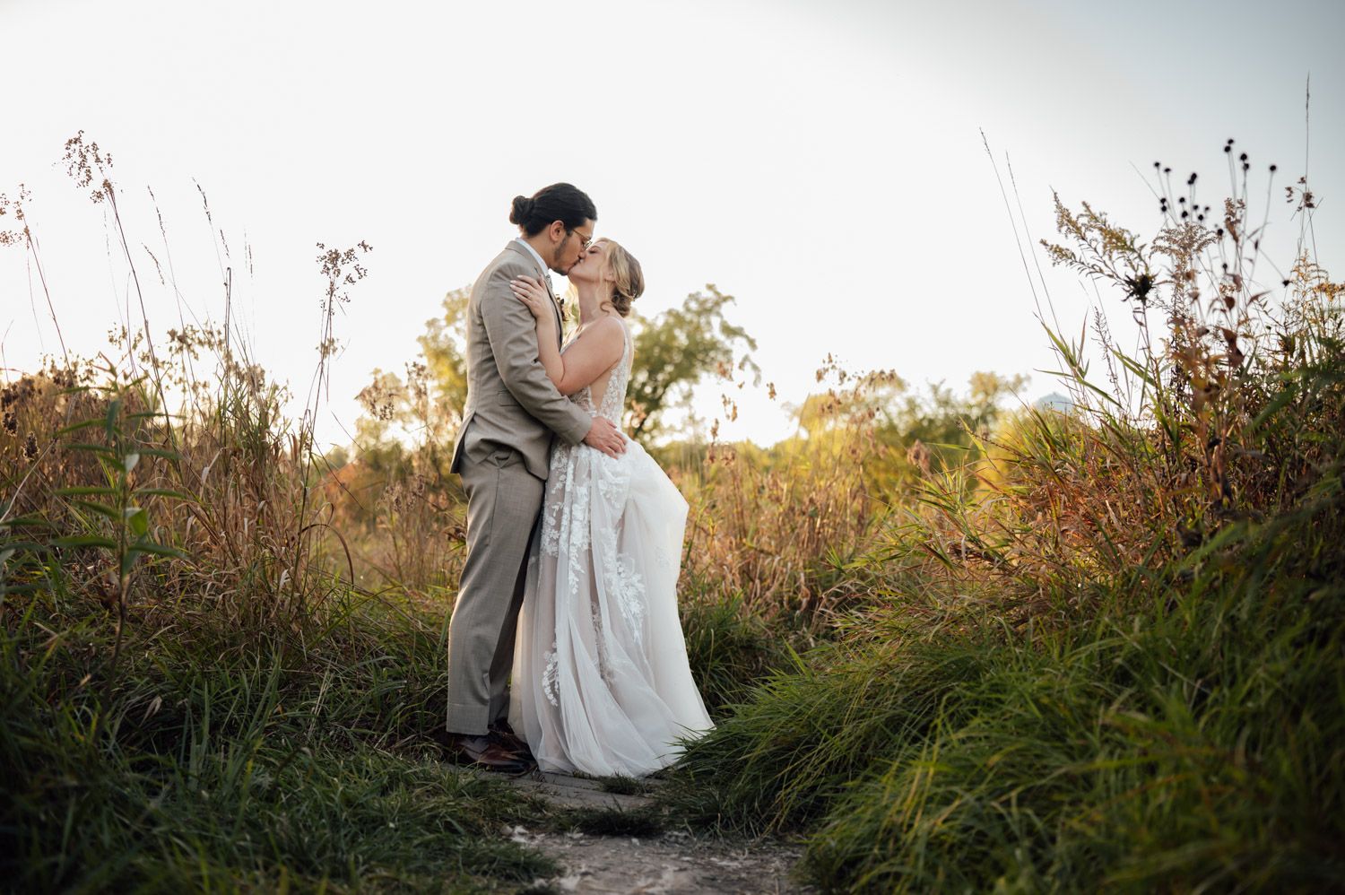 Newlyweds kissing on a nature trail path surrounded by lush green and golden foliage.