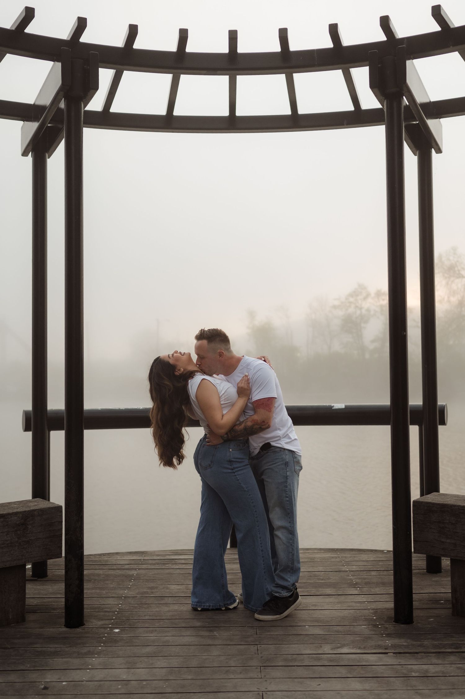 Couple sharing a passionate kiss under a wooden pergola on a misty lakeside dock.