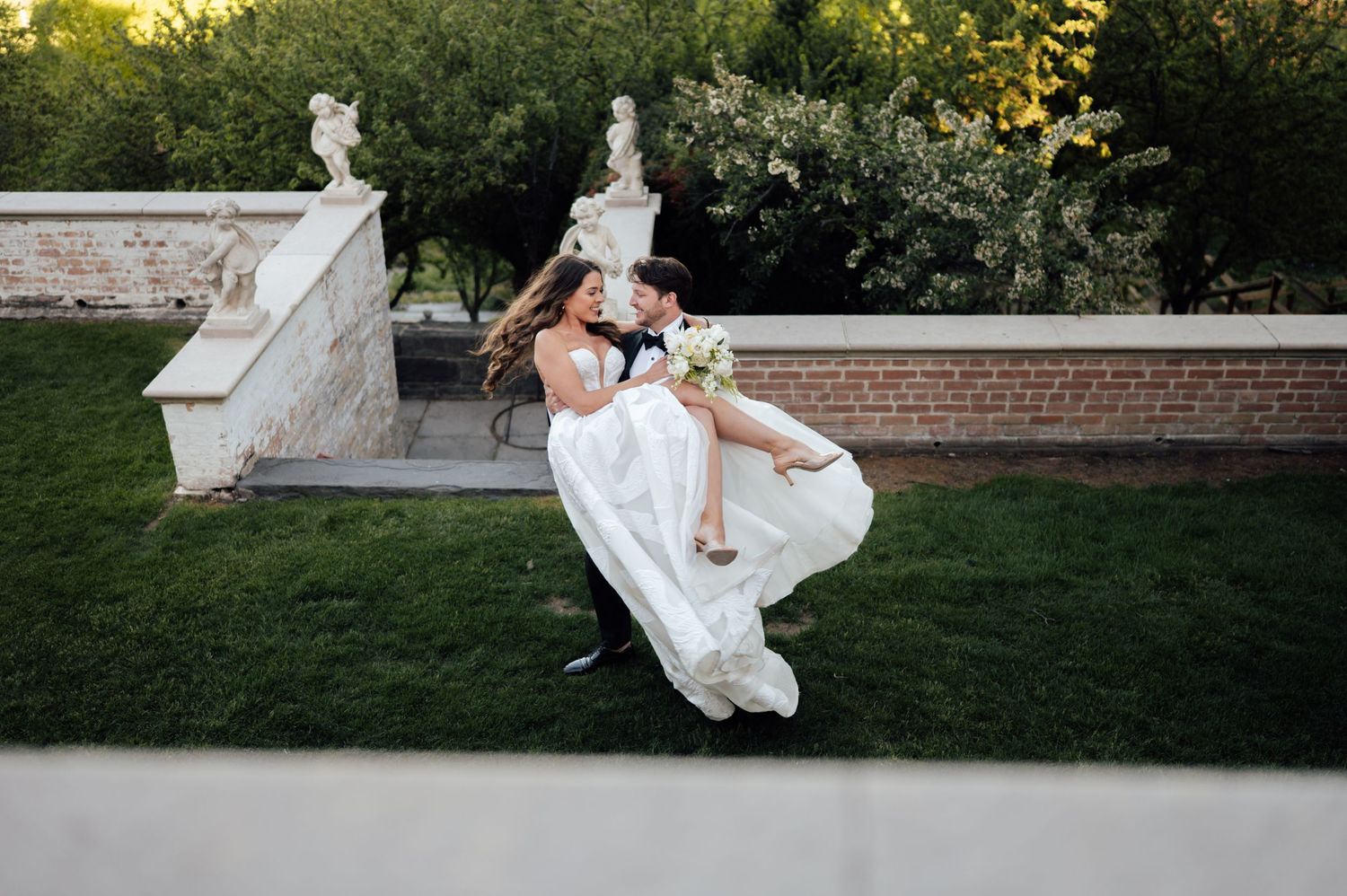 Groom lifts bride in white gown outdoors near stone wall and garden statues at wedding celebration.