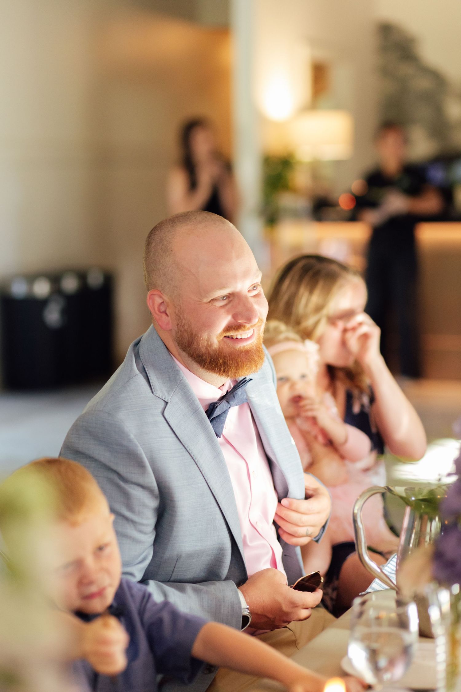 Smiling bald man with red beard in gray suit at wedding reception dinner table with guests in background.