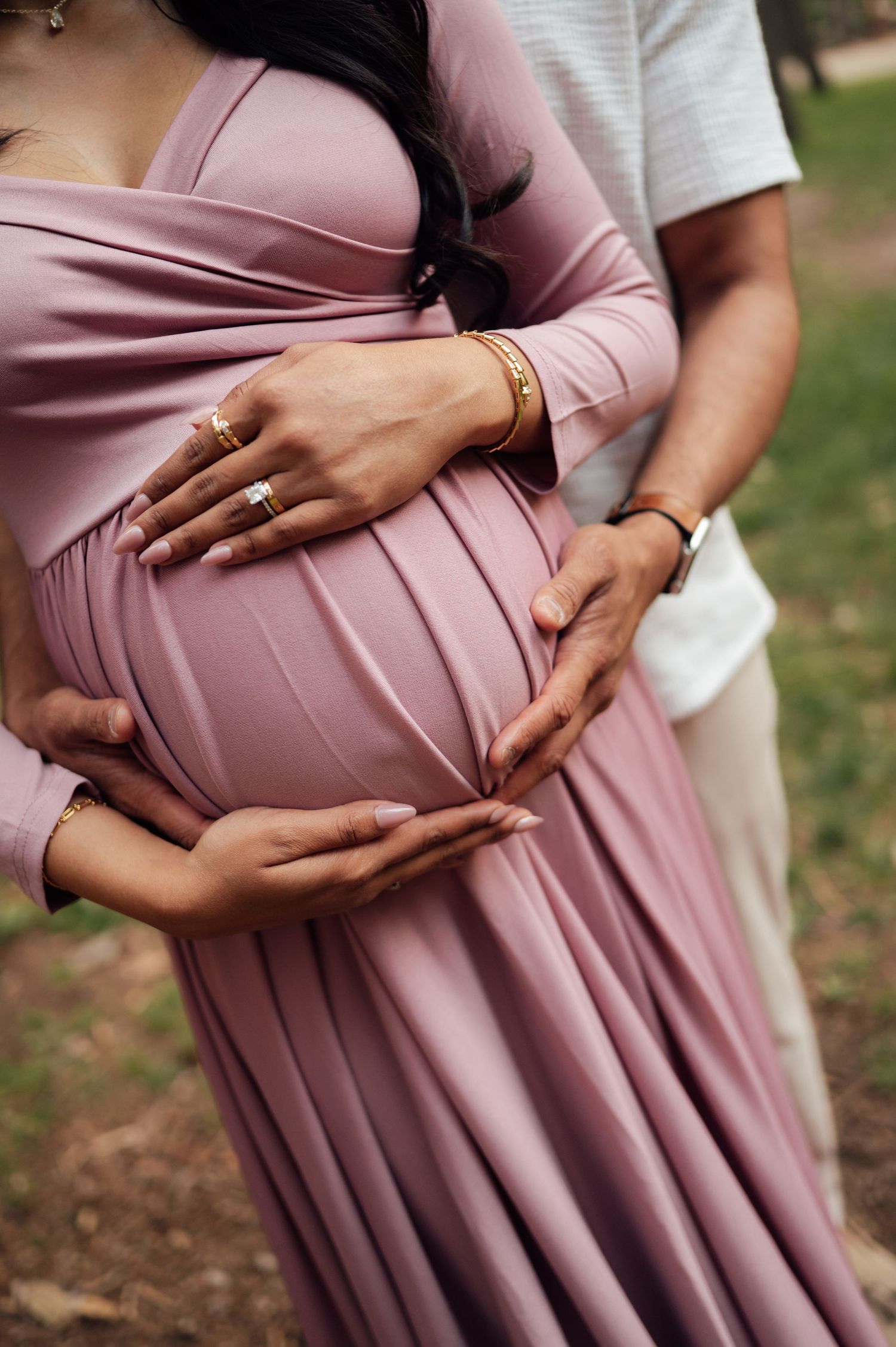 Pregnant woman in pink mauve dress with partner's hands cradling baby bump during outdoor maternity photoshoot.