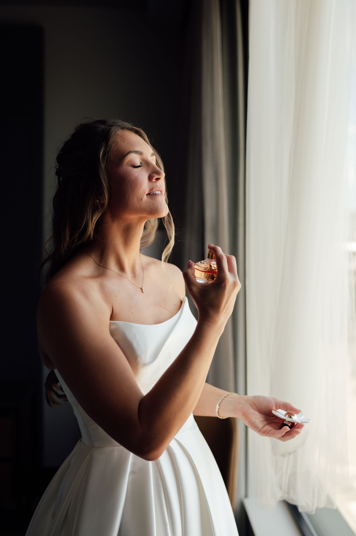 Bride in white strapless gown applying perfume by a window, eyes closed, with soft natural light.