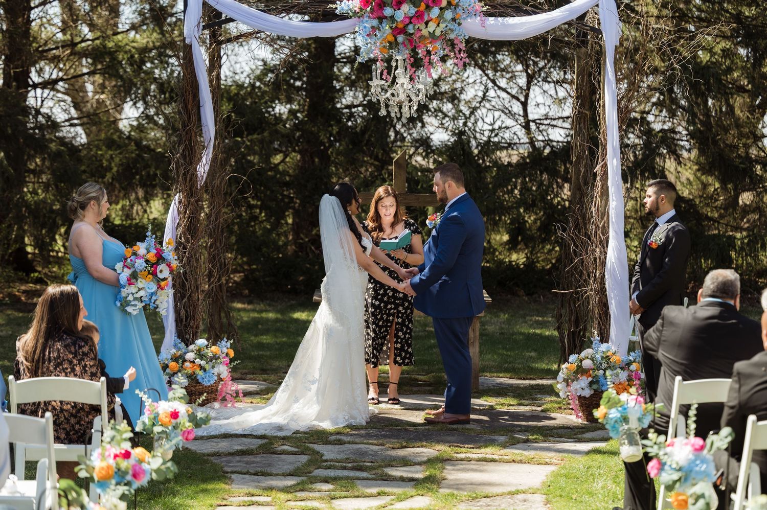 Outdoor wedding ceremony in a garden setting with rustic wooden arch decorated with white fabric and pink flowers.
