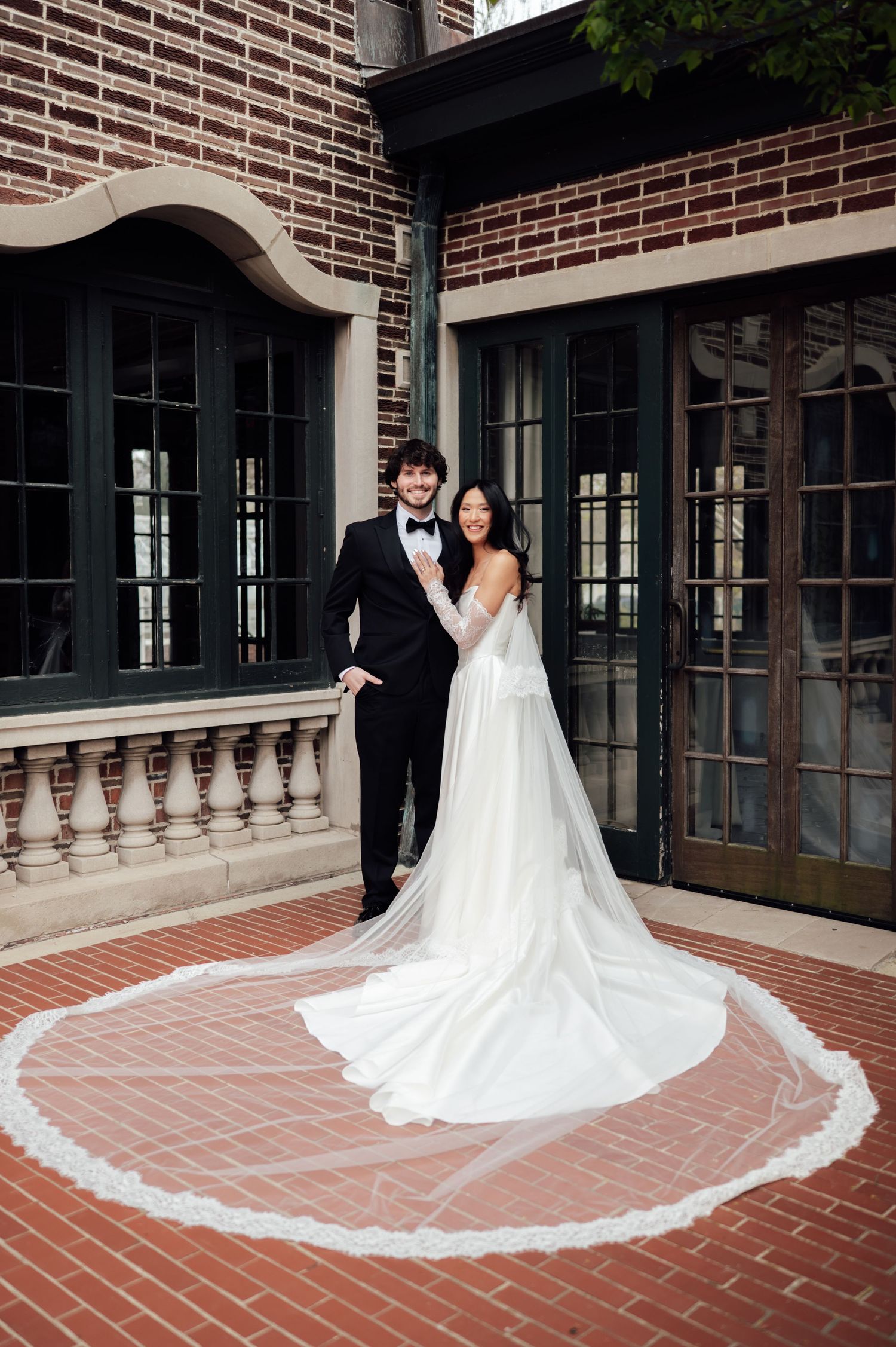 A bride in a long white gown with cathedral veil and groom in black tuxedo pose on a brick terrace.