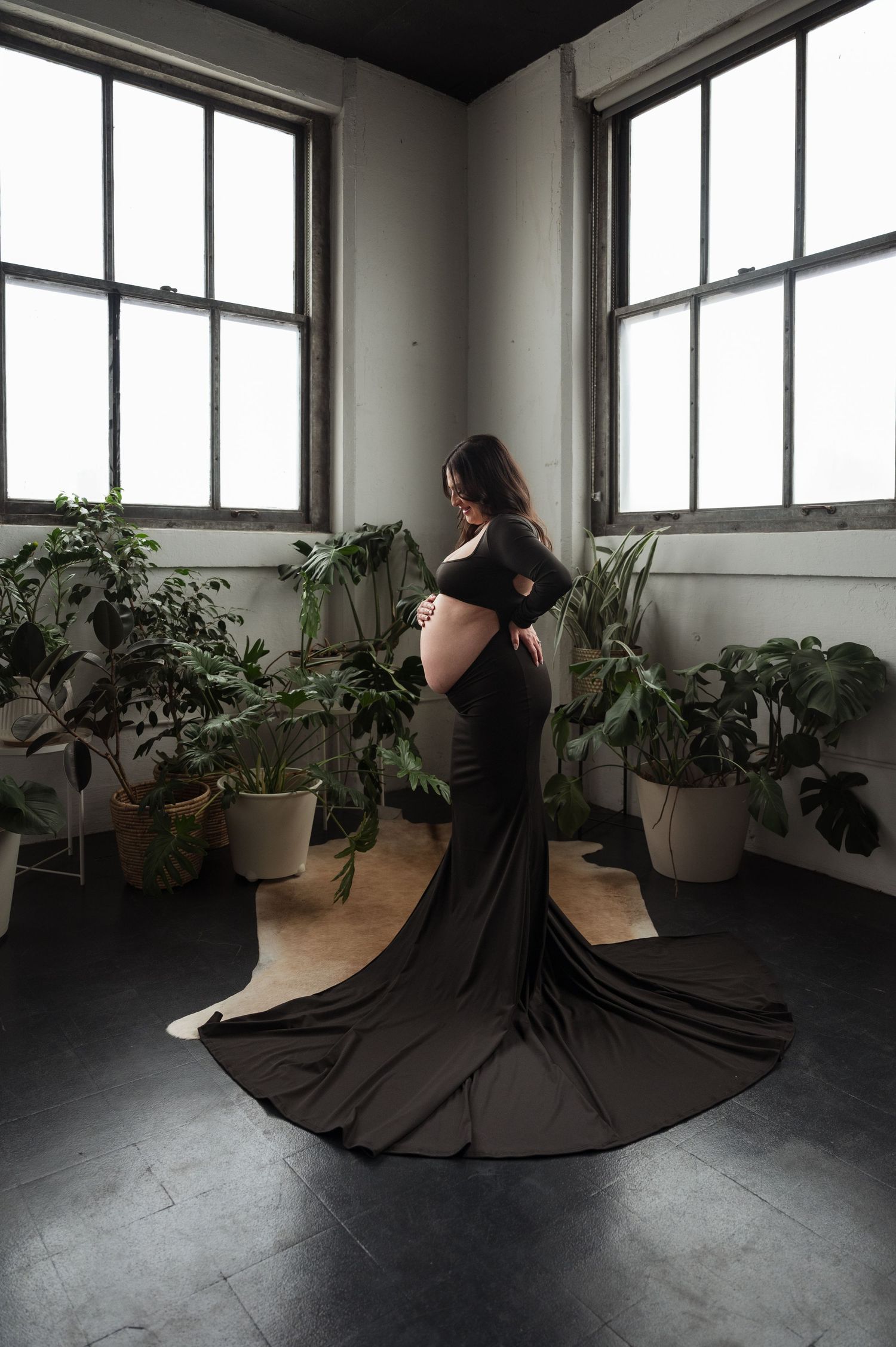 Pregnant woman in black gown with train stands among potted plants by windows.