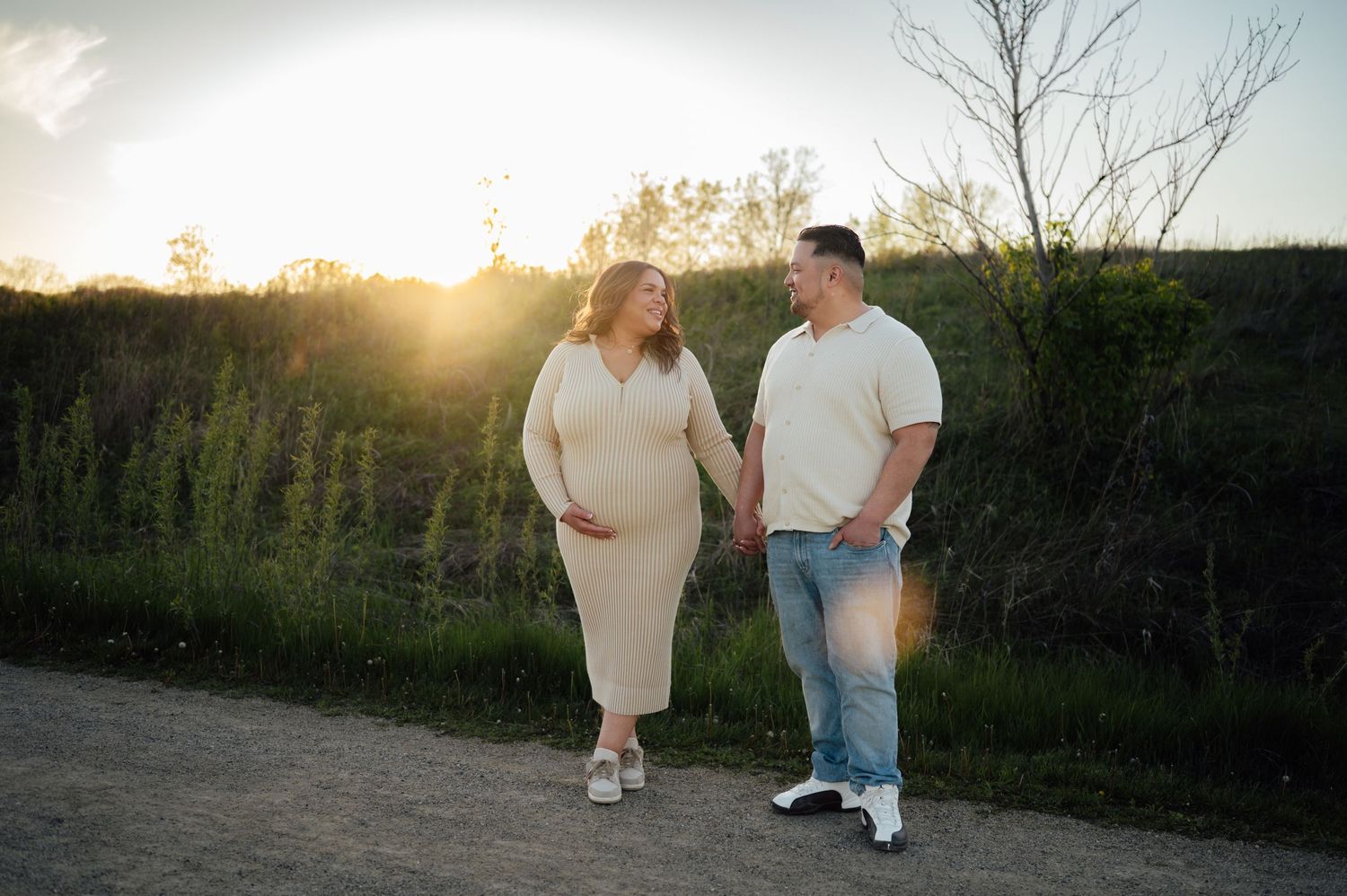 Pregnant couple holding hands walking on a path at sunset with wildflowers behind them.