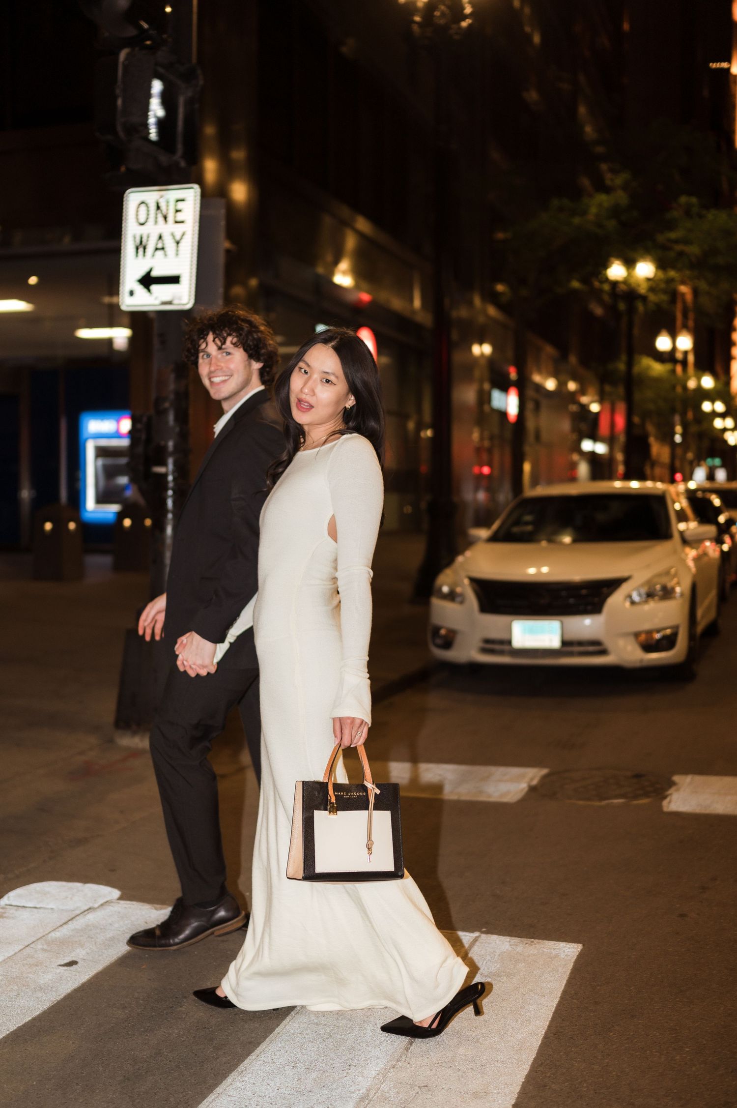 Stylish bride and groom posing back-to-back at a Chicago crosswalk at night near a one-way street sign.