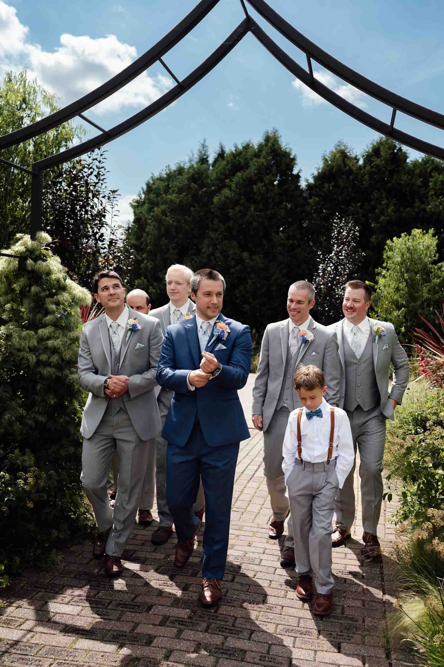 Groom in blue suit leads groomsmen in gray suits along brick garden path at outdoor wedding ceremony.