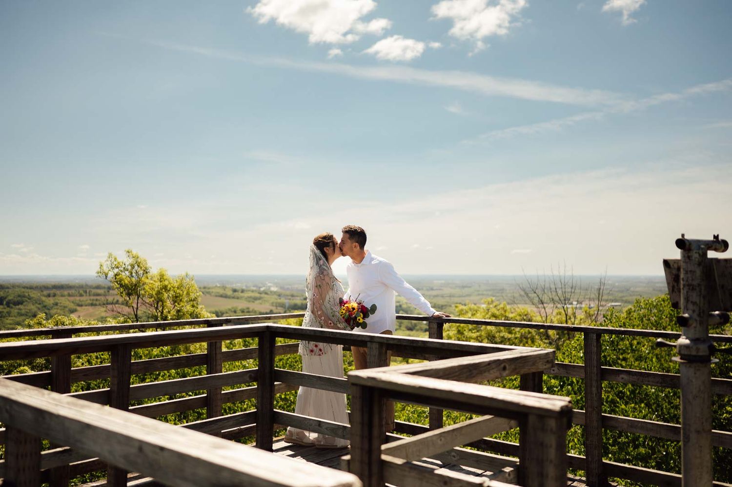 Bride and groom kissing on wooden overlook deck with scenic green landscape behind them.