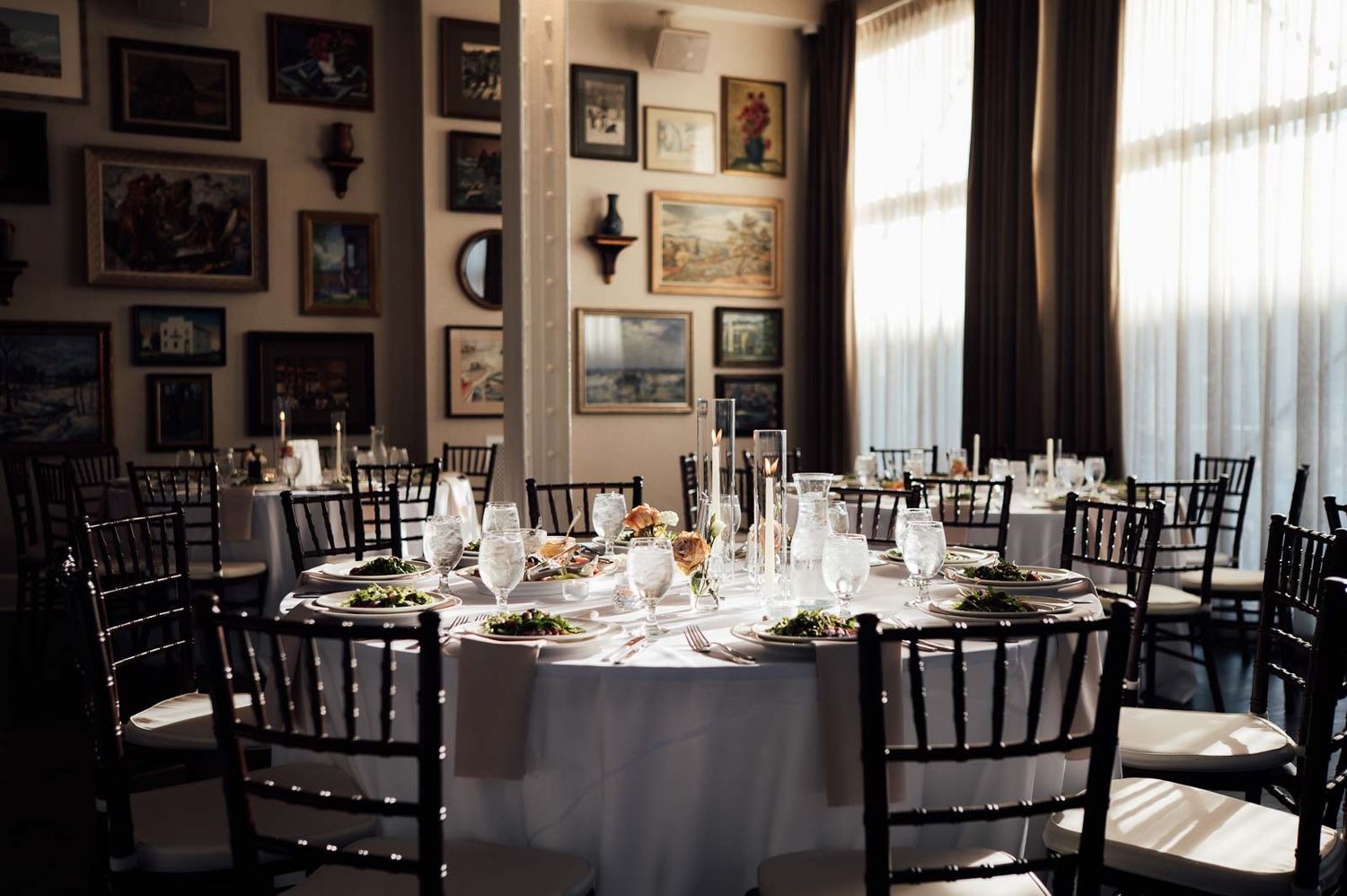 Elegant wedding reception table with white linens, black Chiavari chairs, and art-covered walls in a sophisticated venue.