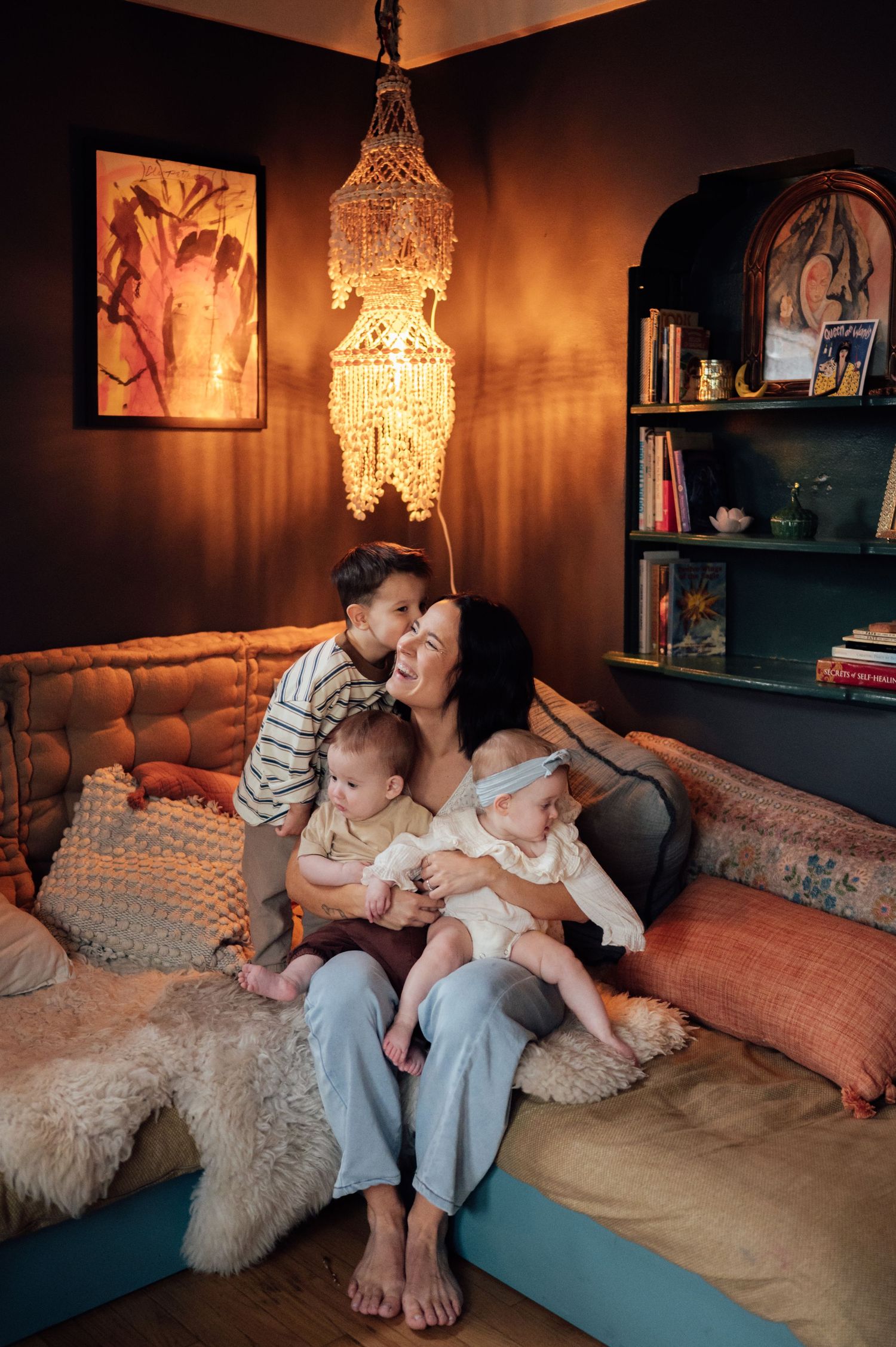 A smiling mother sits on a cozy couch holding twin babies while her young son affectionately hugs her.