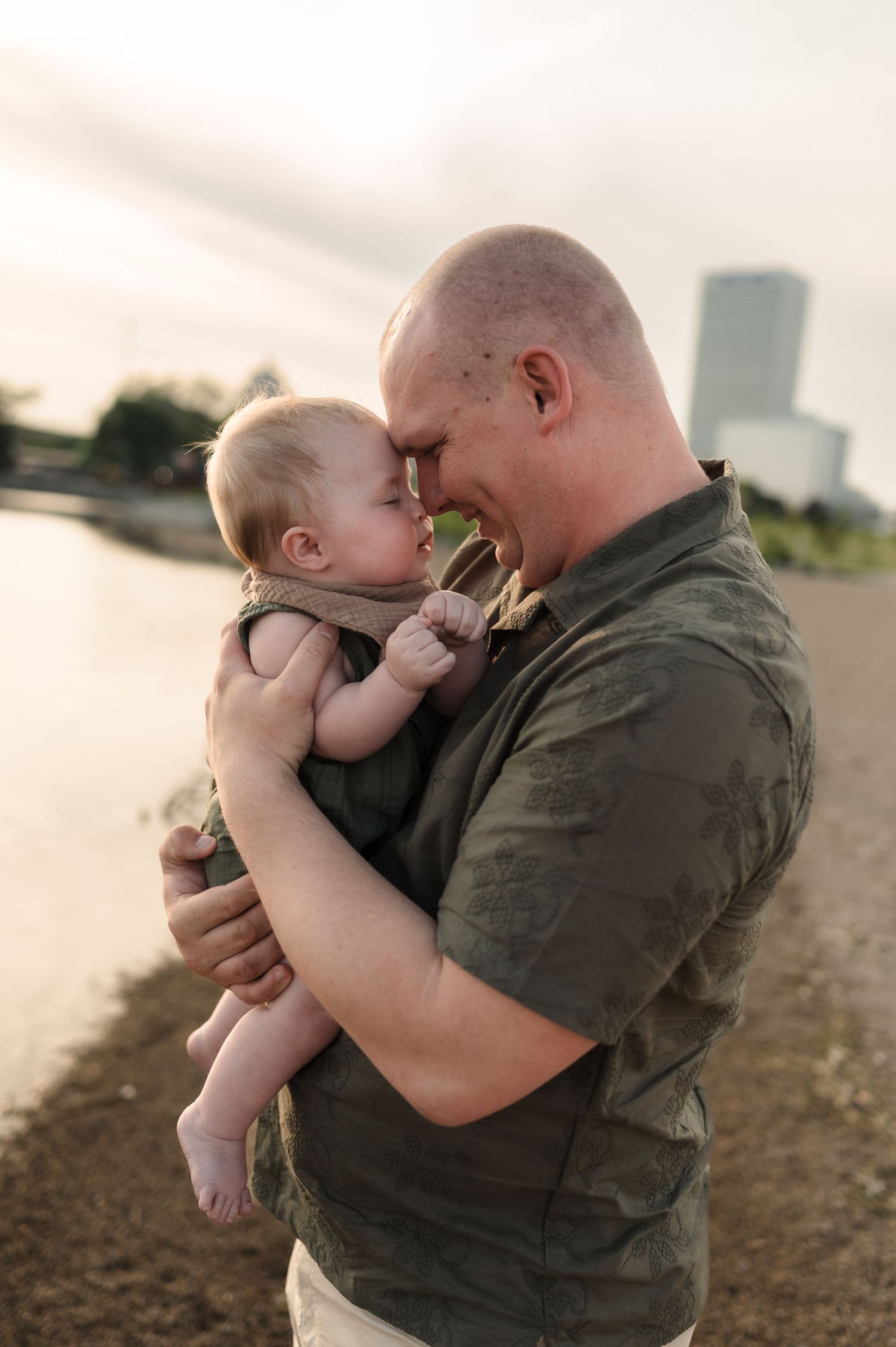 Bald father tenderly holding his baby face-to-face on a sandy beach with city skyline behind.