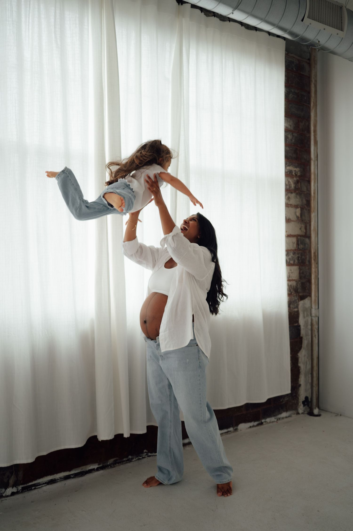 Pregnant woman lifting young child in the air near sheer white curtains in bright airy room.