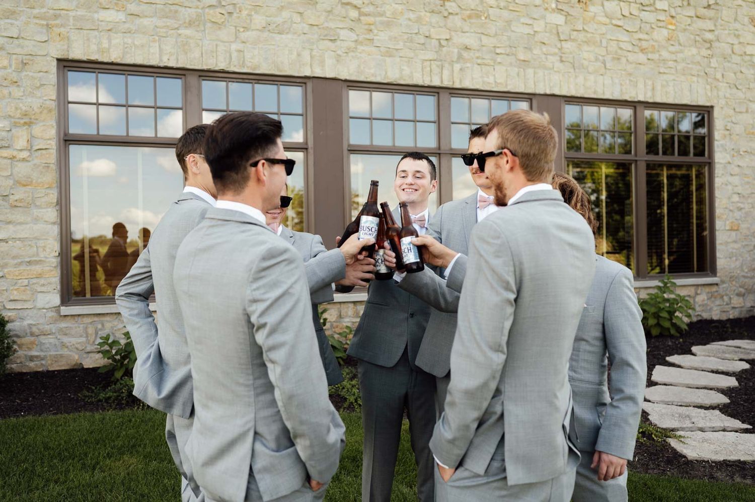 Groomsmen in light gray suits toasting with beer bottles outside a modern stone venue.