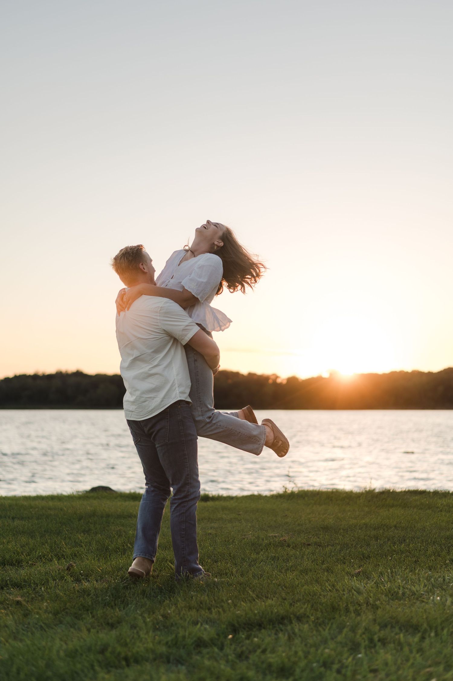 Man joyfully lifts woman in his arms on grassy lakeside lawn during a vibrant golden sunset.