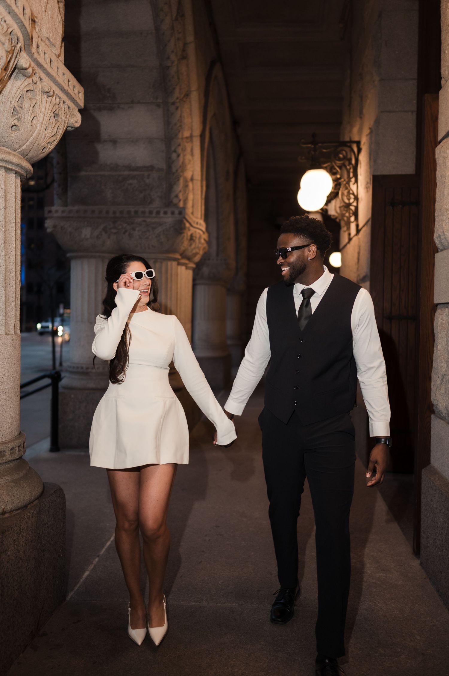 An elegant couple in white dress and black vest hold hands while walking through an ornate building at night.
