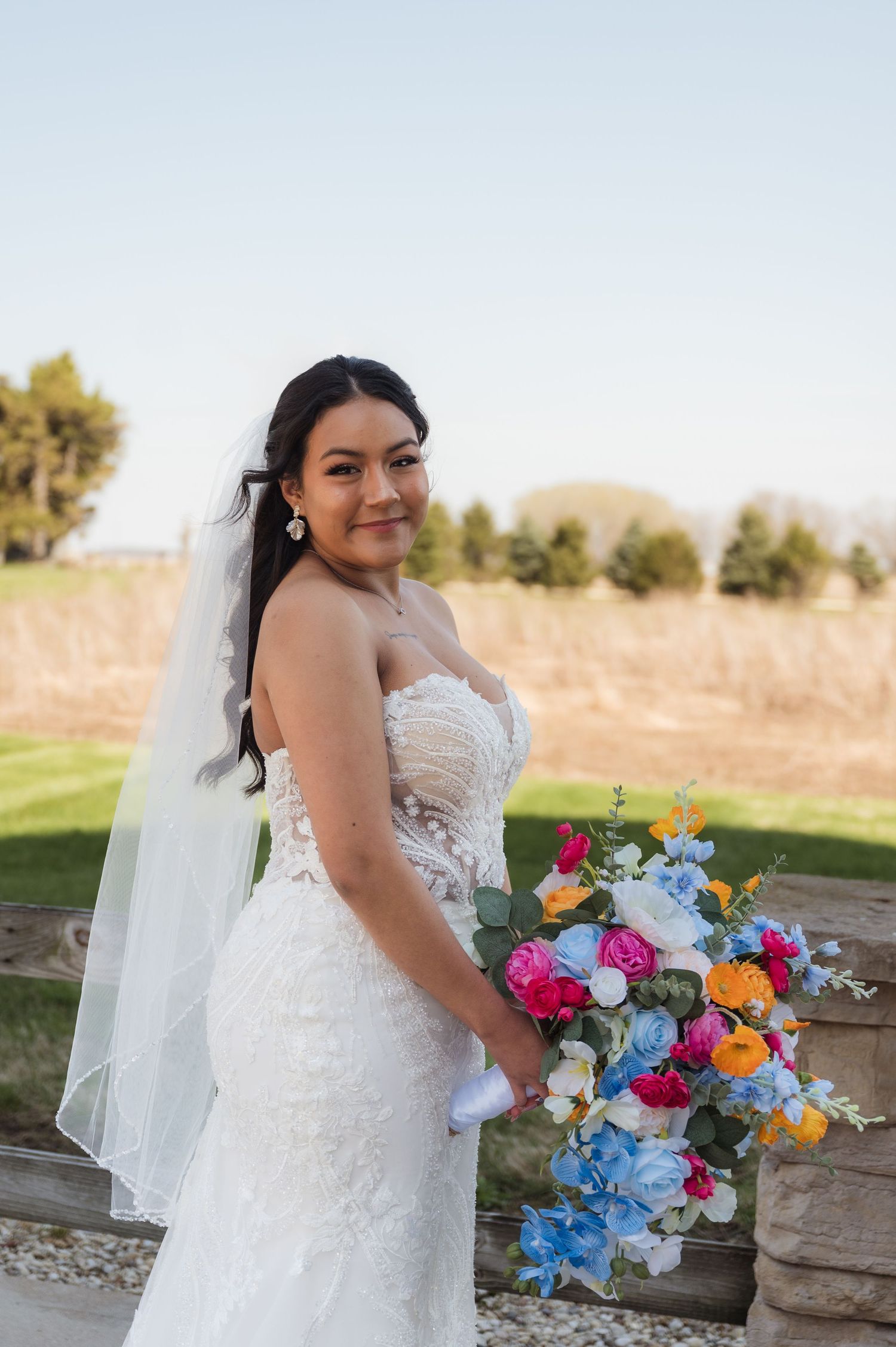 Bride in a lace strapless gown holds vibrant multicolored bouquet with blue and pink flowers at outdoor venue.