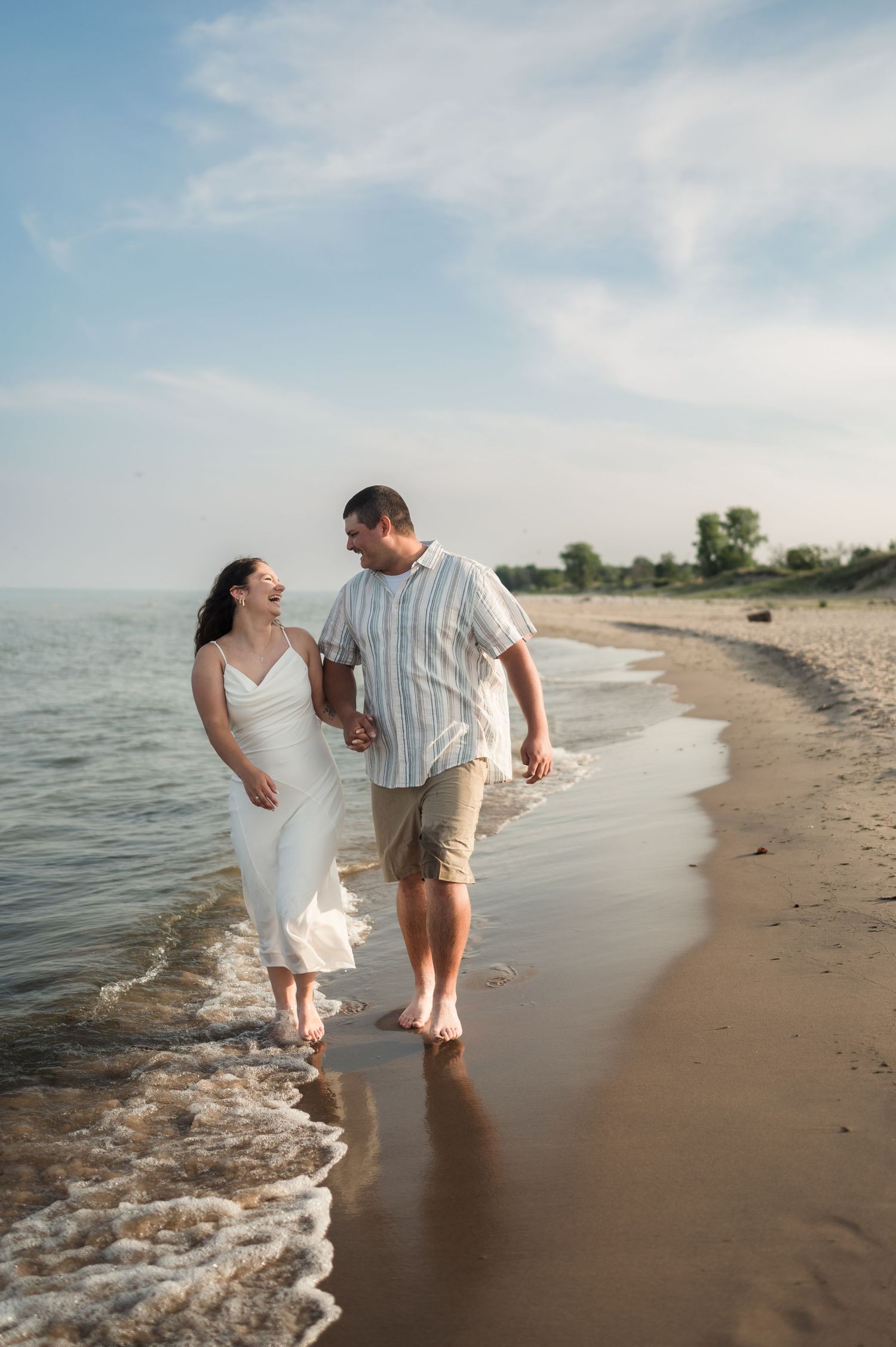 A couple walks barefoot along a serene beach shoreline on a sunny summer day, waves gently lapping at their feet.