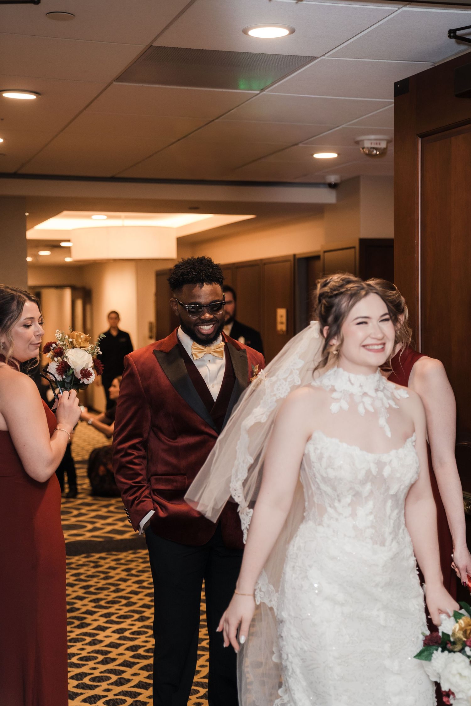 A bride in a white lace wedding dress dances and celebrates in a carpeted hotel hallway.