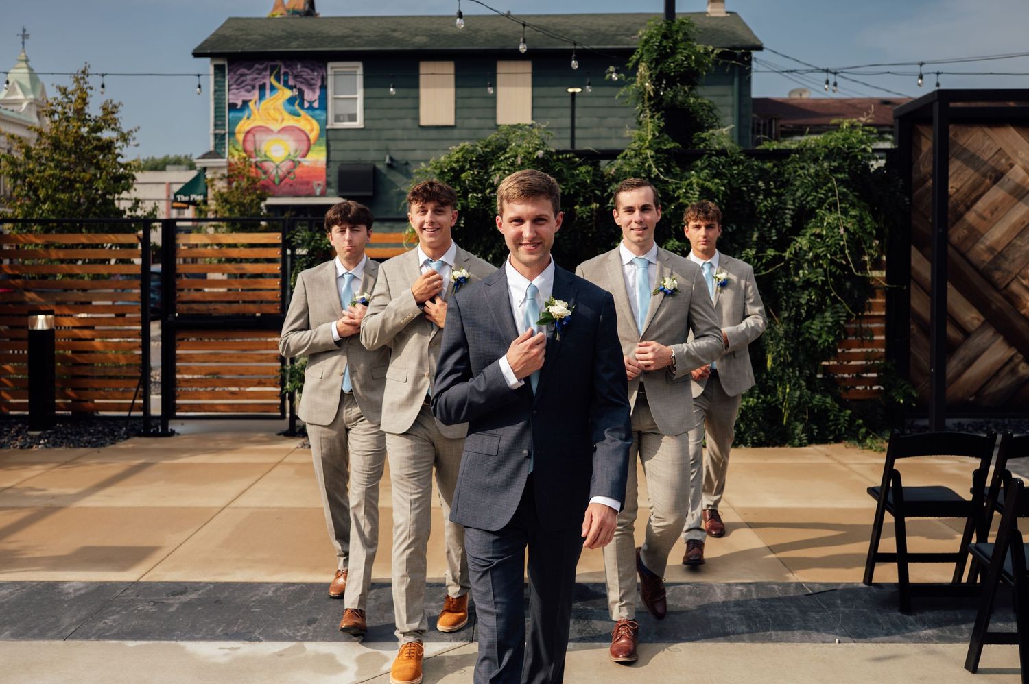 Groom in navy suit leads groomsmen in tan suits walking outdoors at wedding venue on sunny day.