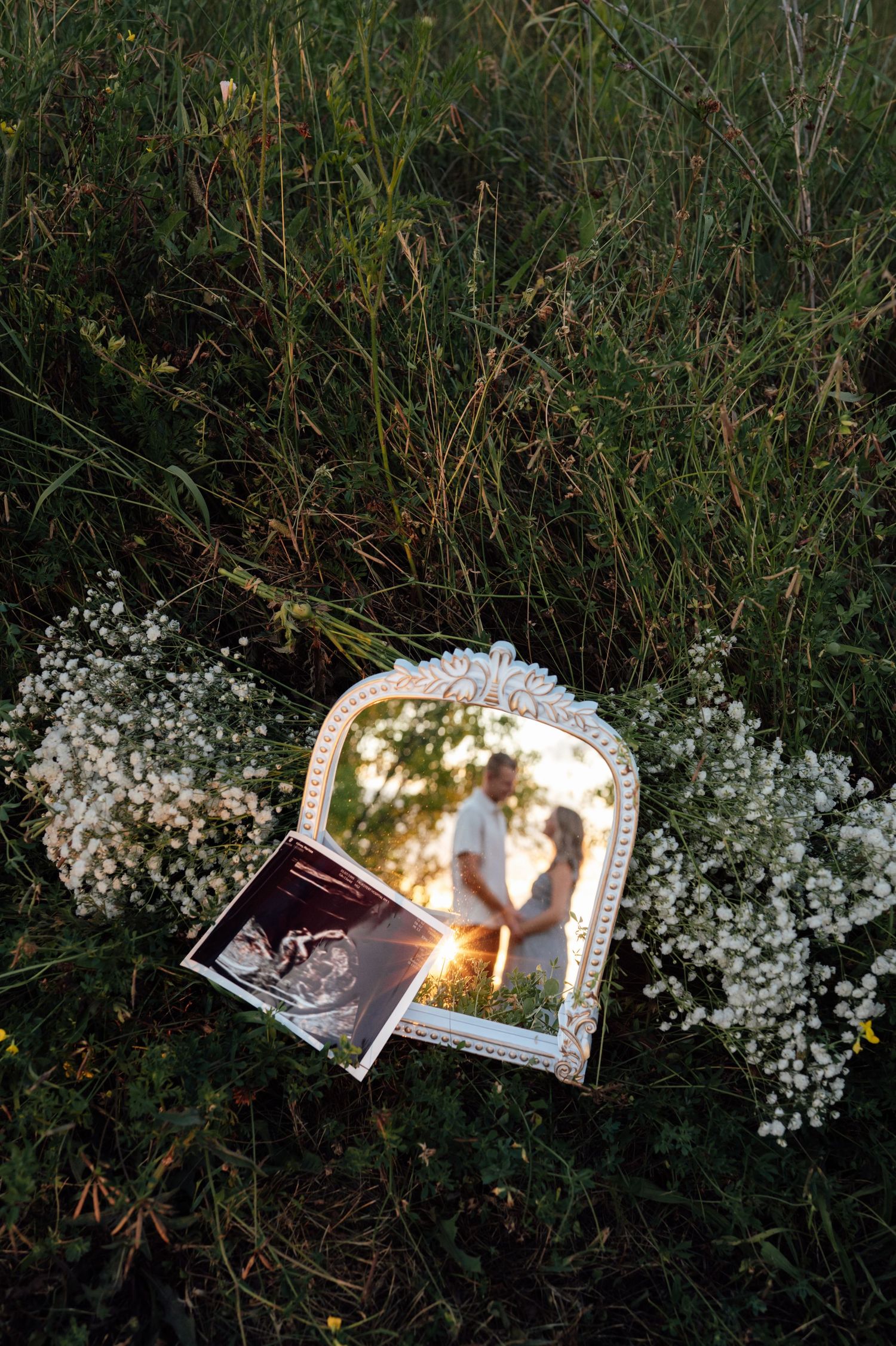 A decorative mirror and ultrasound photo rest on wildflowers in grass, reflecting a couple embracing at golden hour.