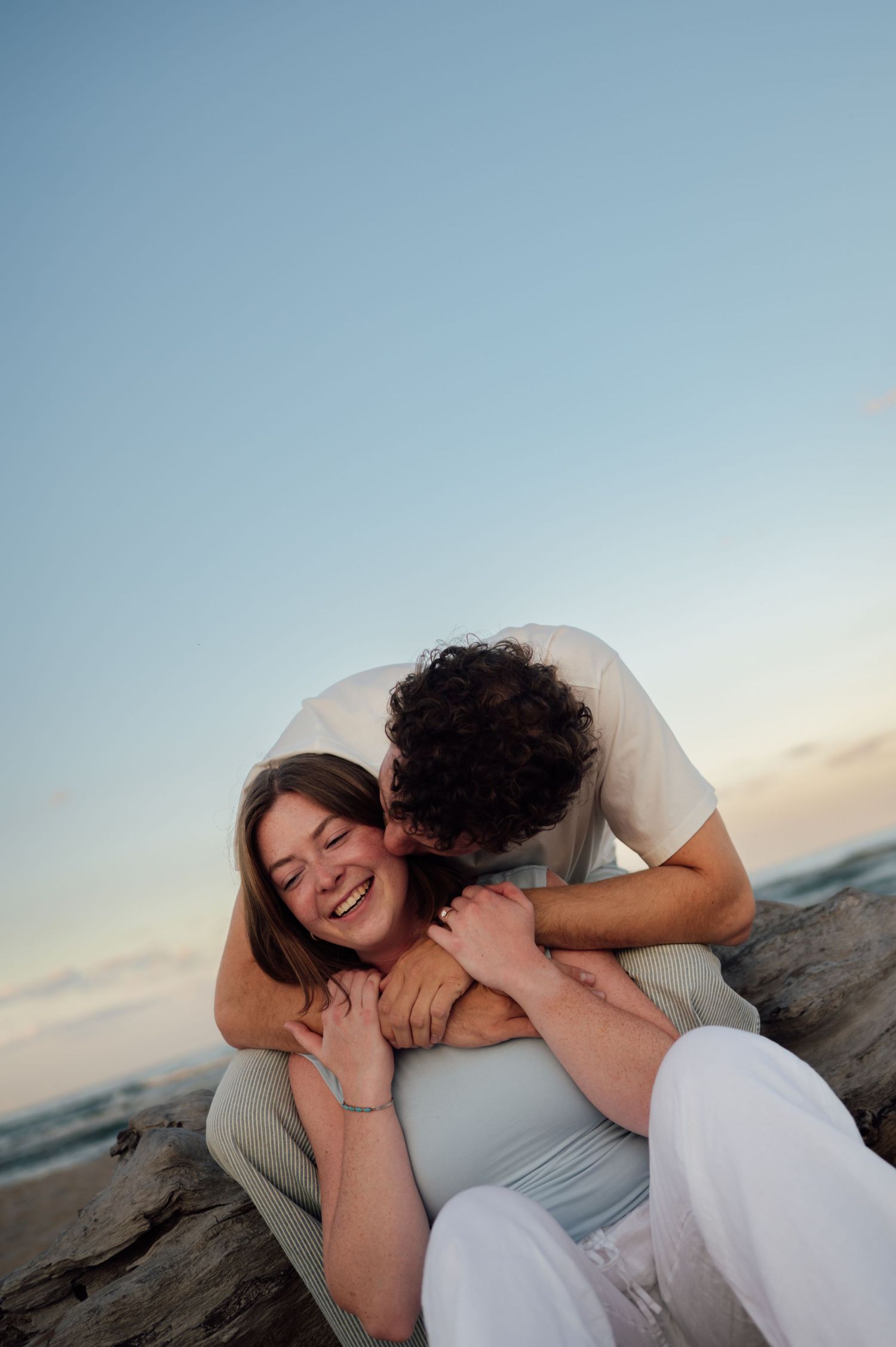 A couple shares a tender moment on a beach at sunset, with the man kissing the laughing woman as she sits between his legs.