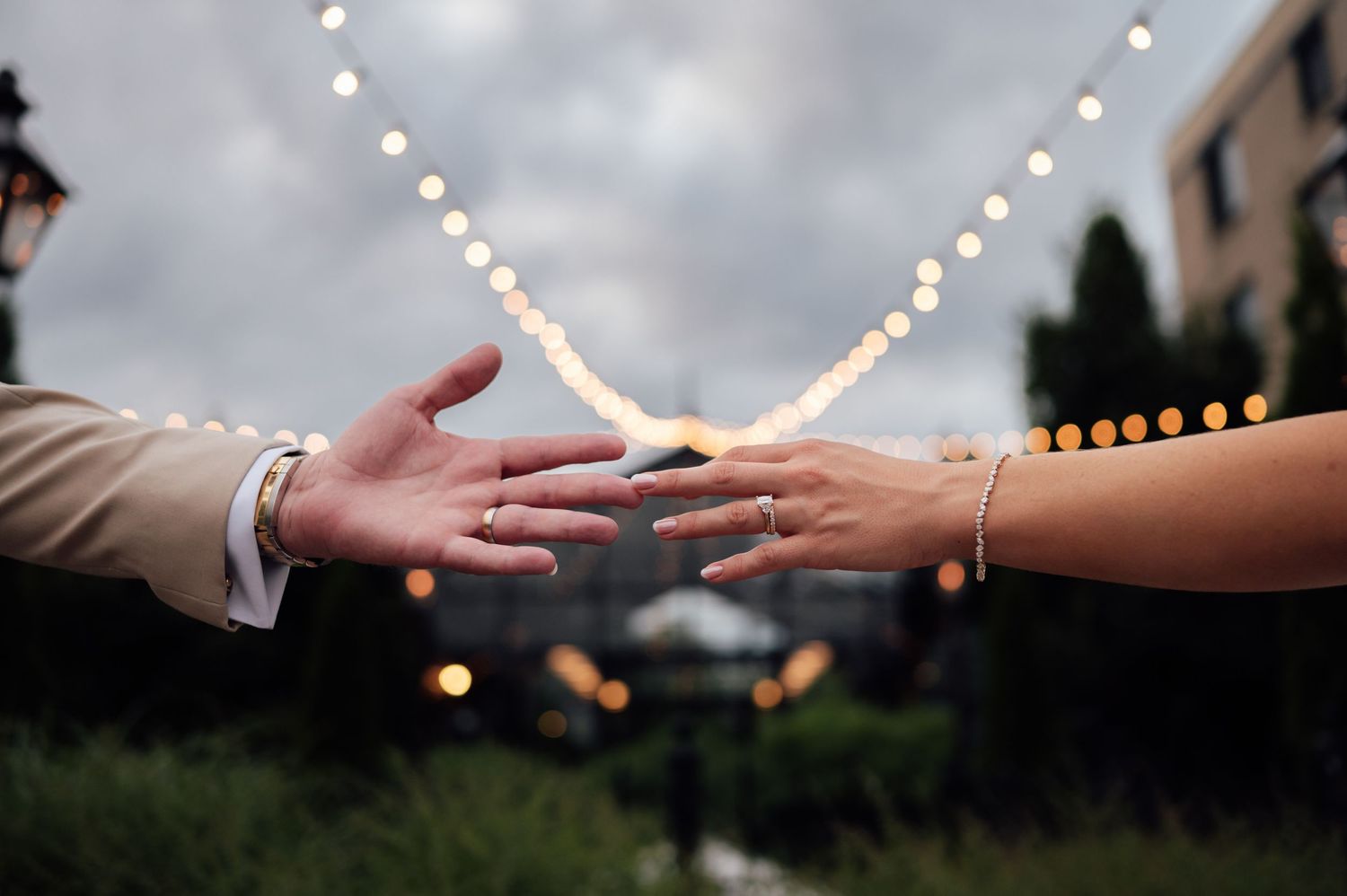 Two hands reaching toward each other with wedding rings, string lights glowing softly in the background at an outdoor venue.