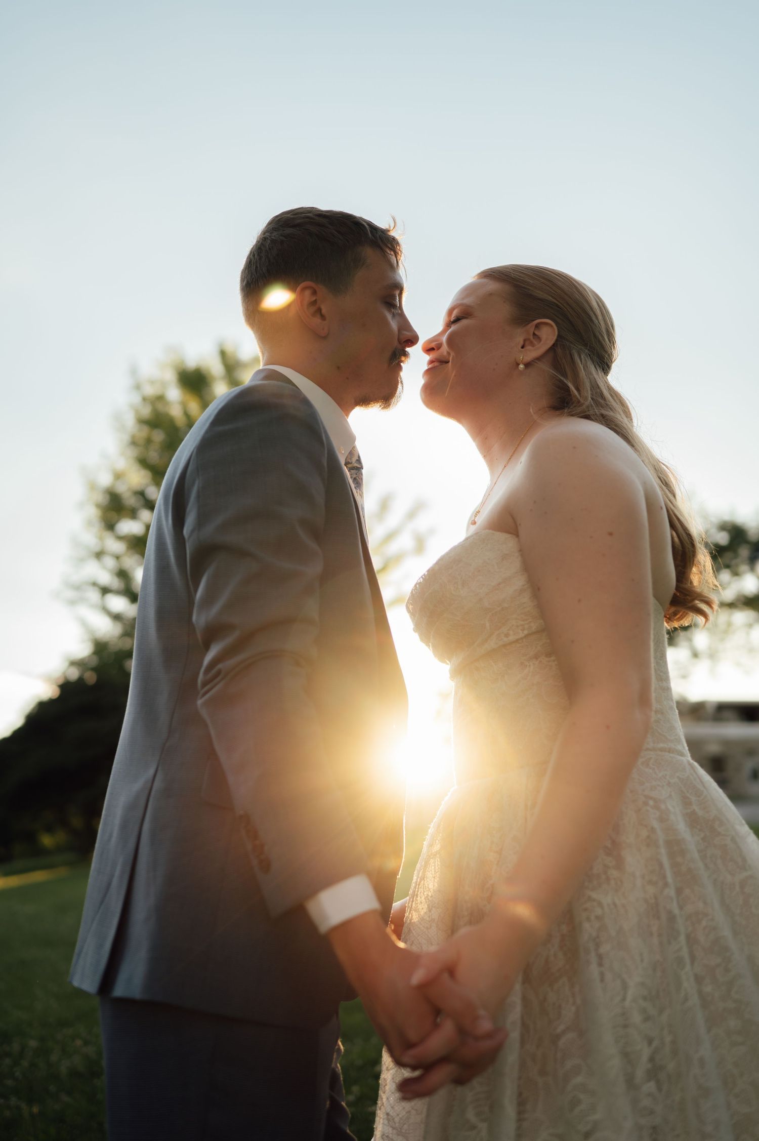 Bride and groom share a romantic sunset kiss while holding hands outdoors on their wedding day.