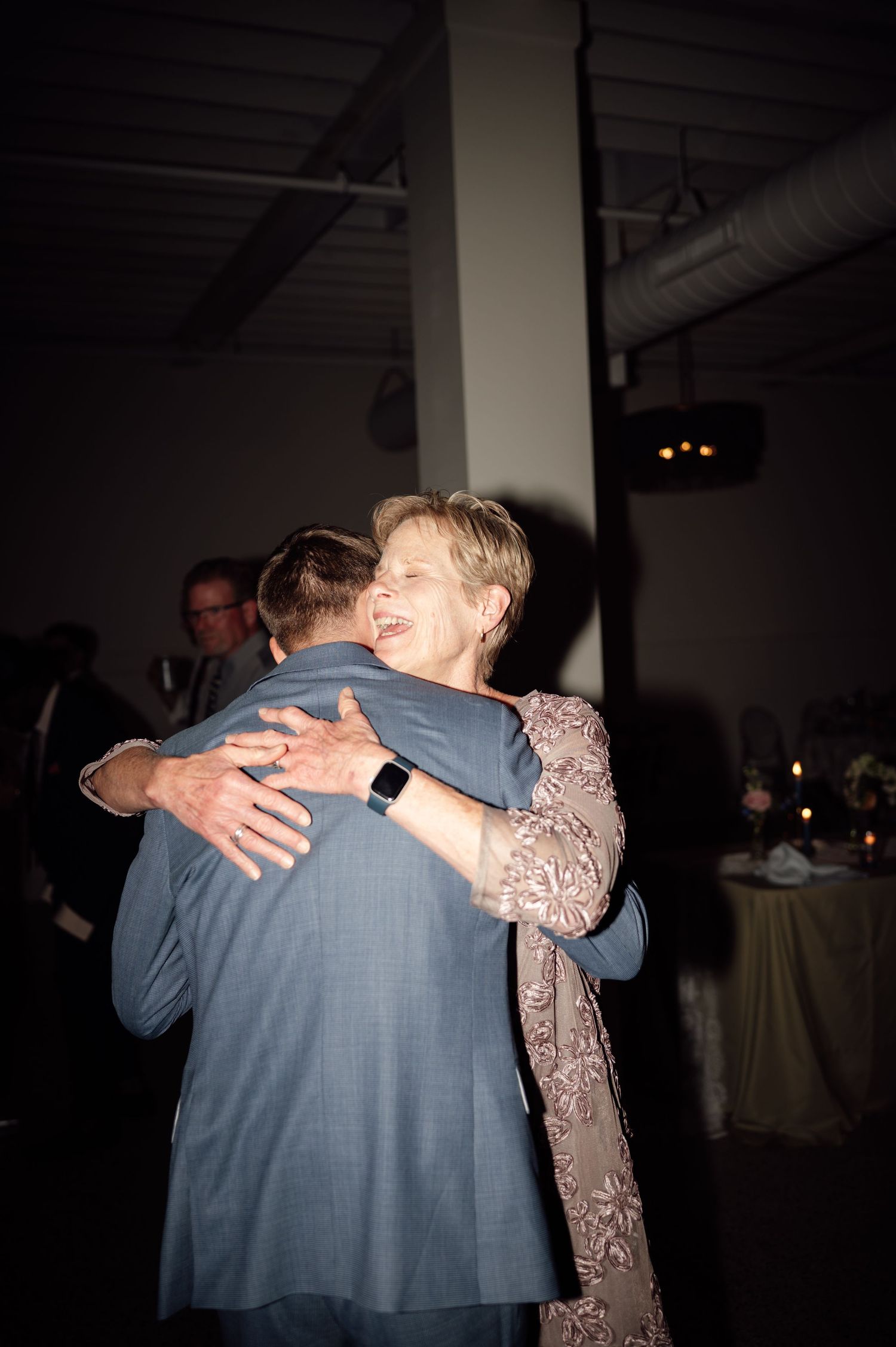 Elderly woman in pink lace dress hugging a man in gray suit at a formal wedding reception event.