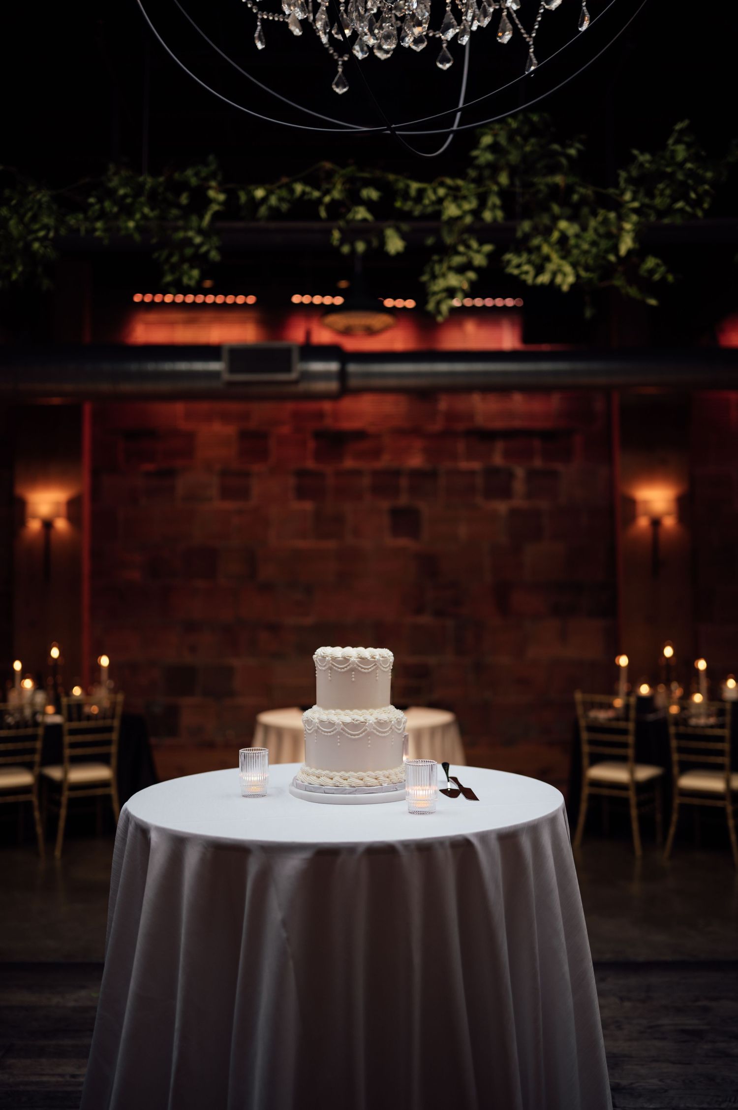 Elegant two-tier white wedding cake on draped table with candles and brick wall backdrop in dimly lit reception venue.