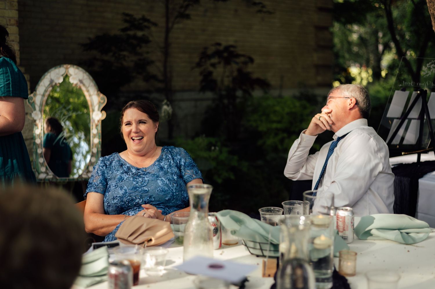 Guests laughing joyfully at an outdoor wedding reception dinner table with greenery in the background.