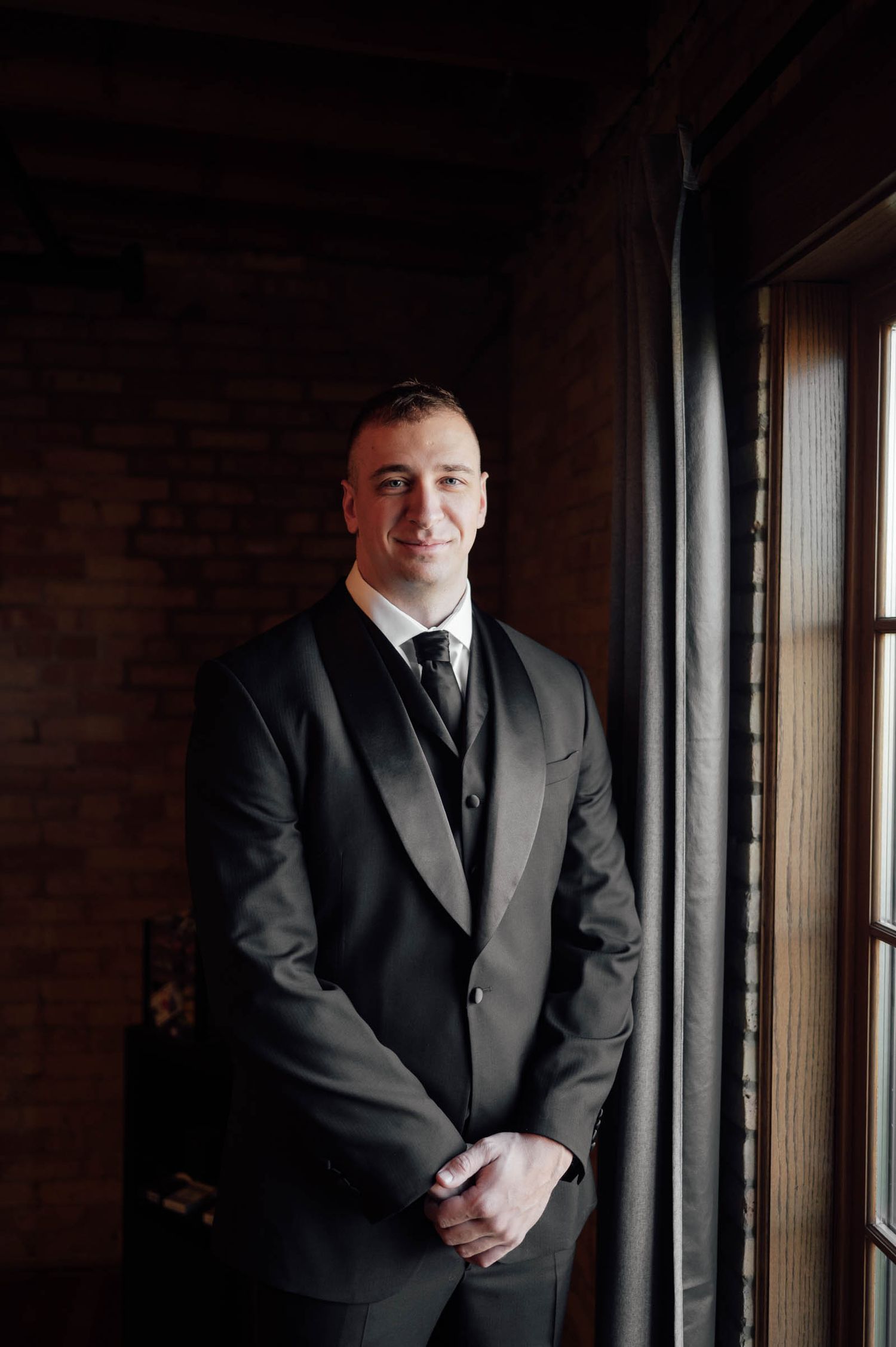 Groom in black tuxedo stands smiling by a window in a rustic brick-walled room.