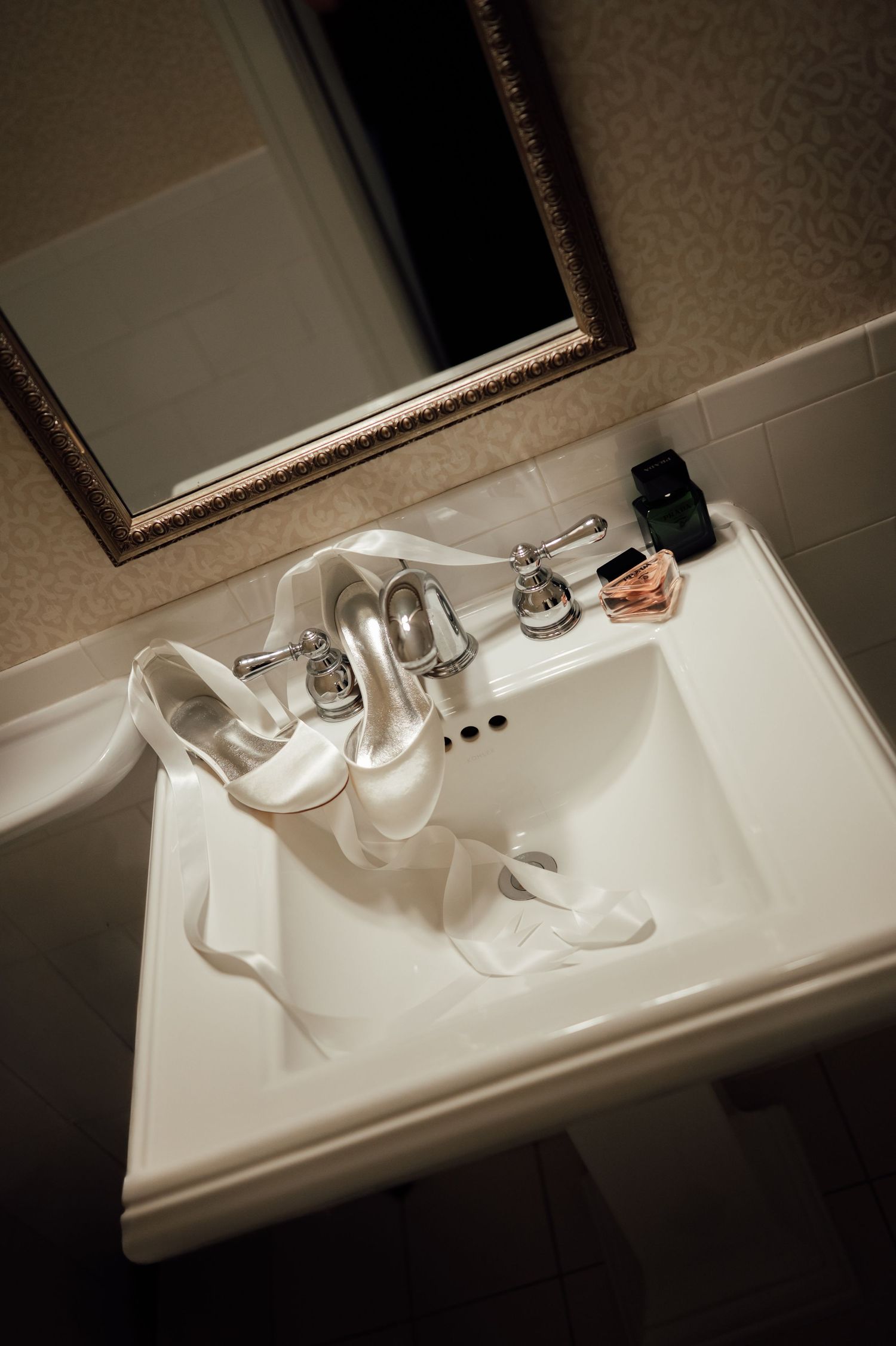 White satin bridal heels rest in a bathroom sink beside perfume bottles and a mirror.
