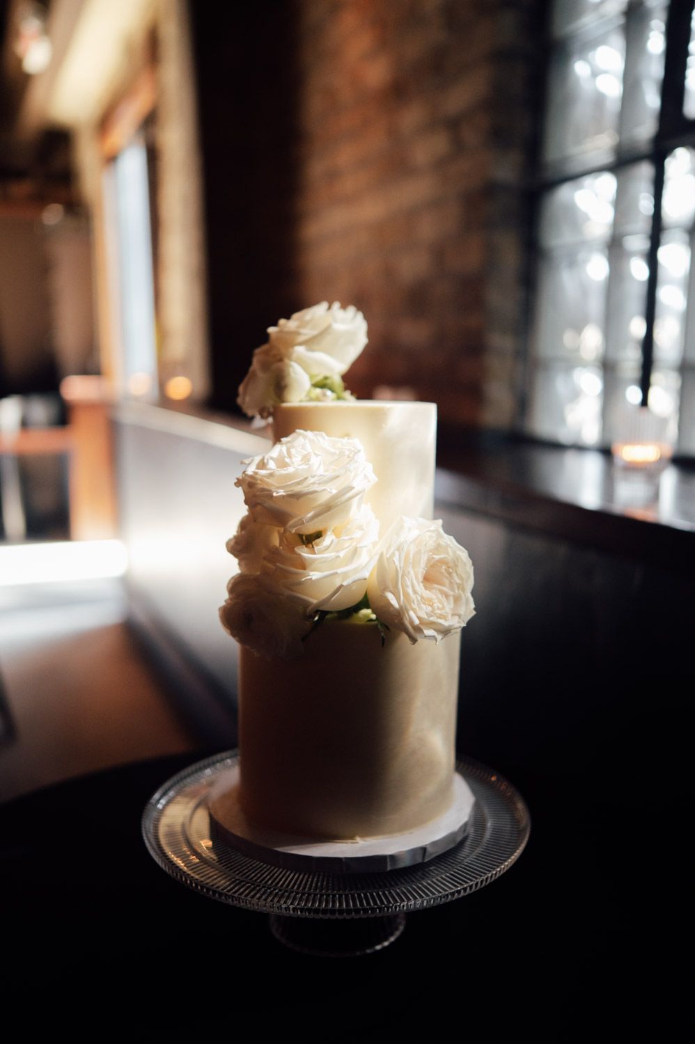 Two-tier white and gold wedding cake decorated with white roses on glass cake stand.