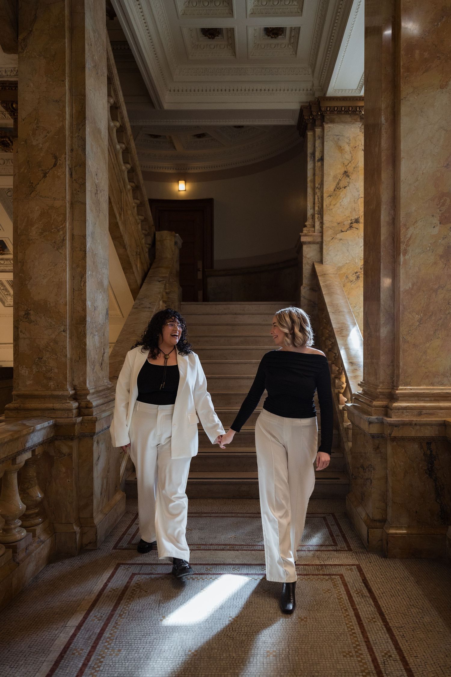 Queer couple in white pants holding hands walking up grand marble staircase together.