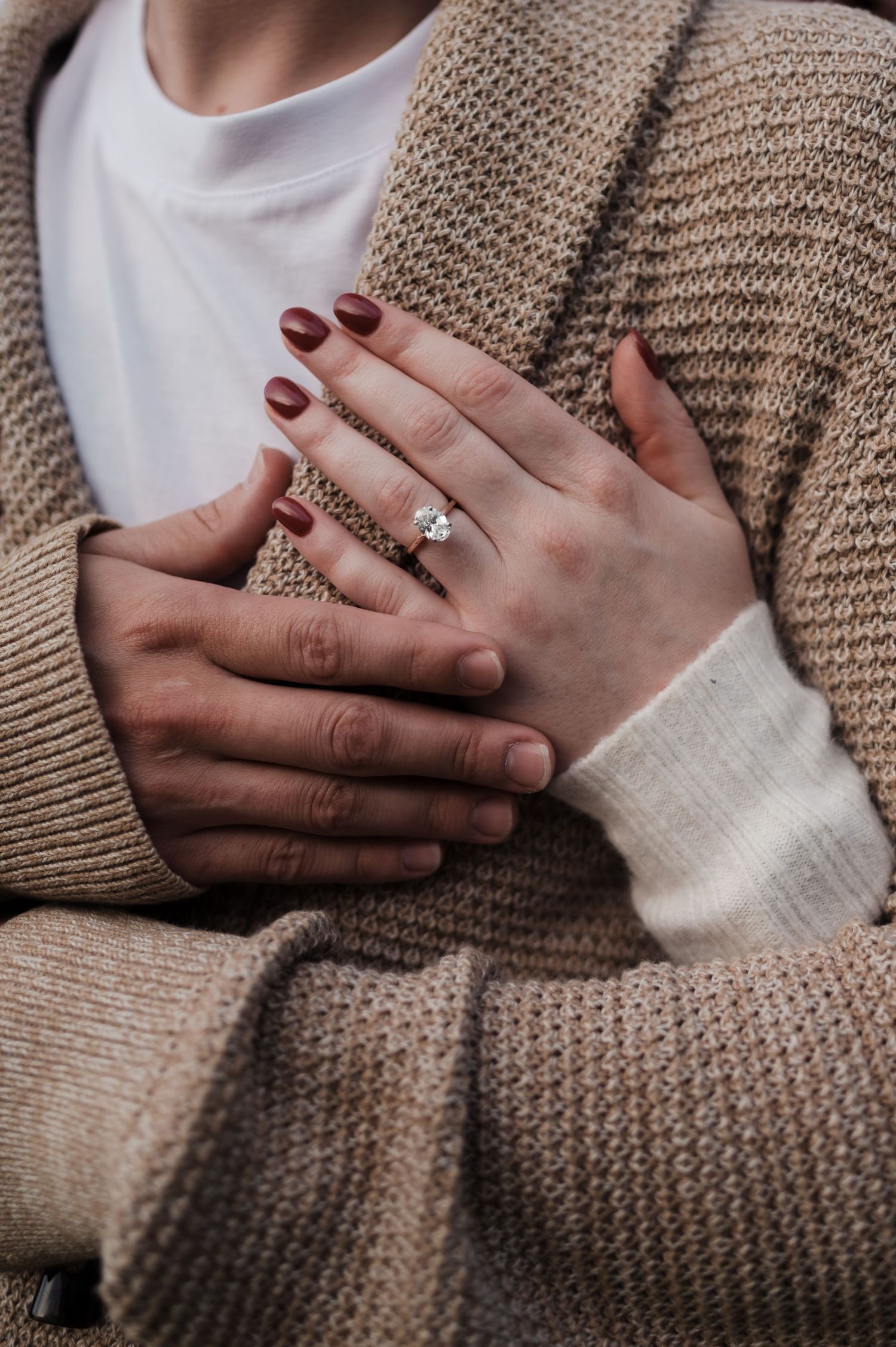 Close-up of woman's hand with diamond engagement ring resting on man's chest in a beige cardigan.