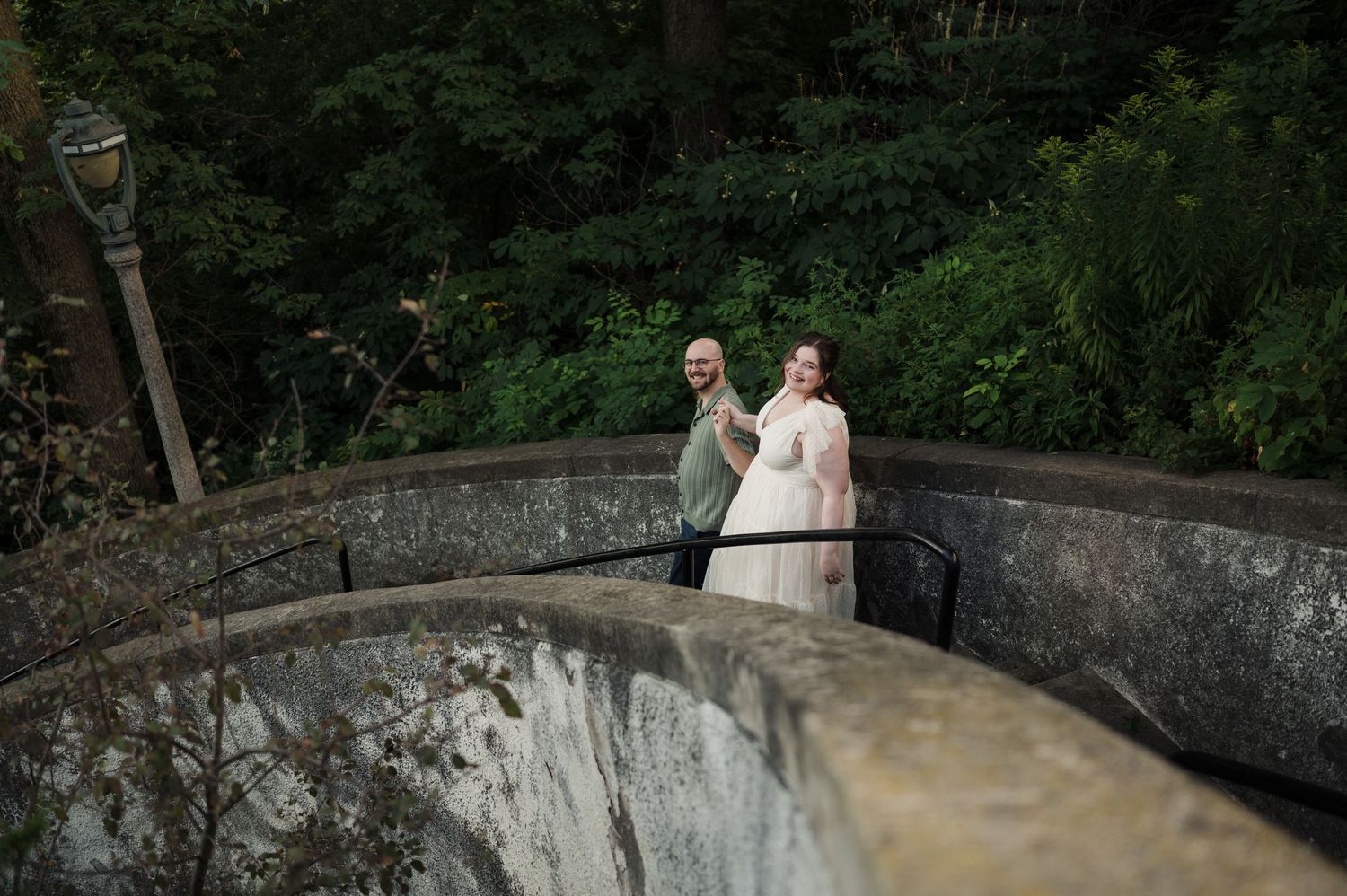 Engaged couple posing on curved stone staircase surrounded by lush green trees during outdoor photo session.