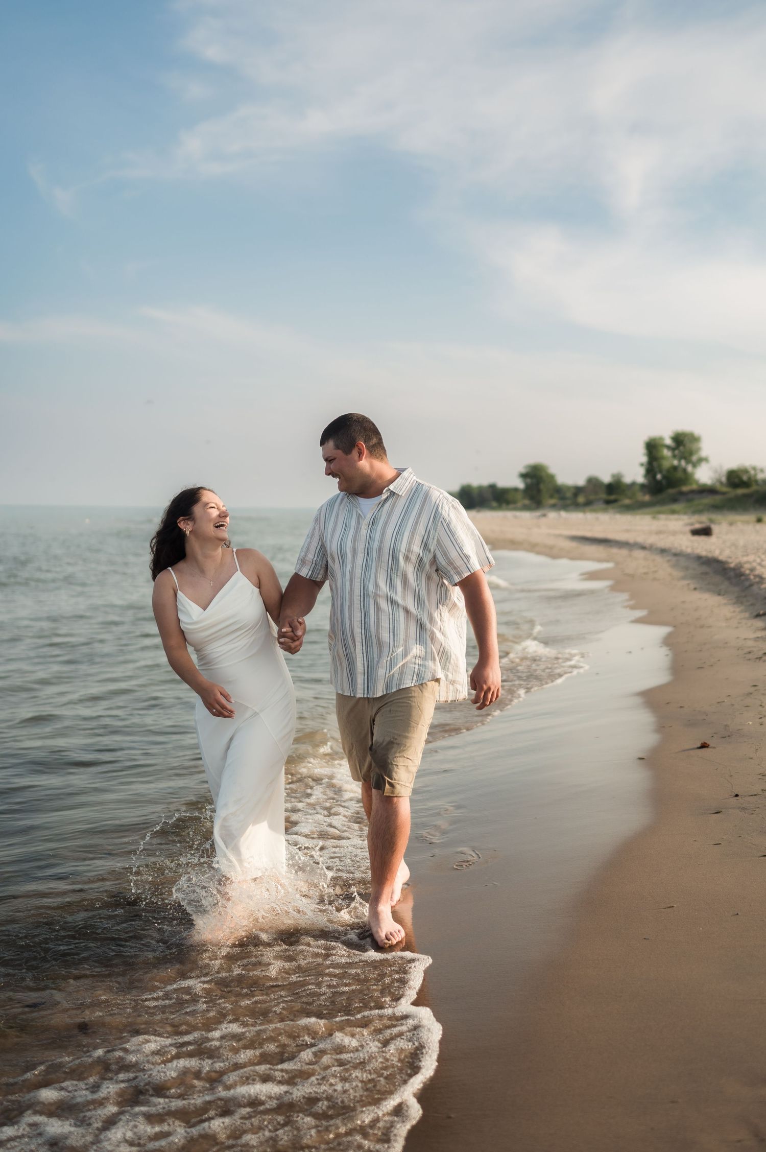 Couple holding hands laughing while walking along sandy beach shoreline with waves splashing around their feet.